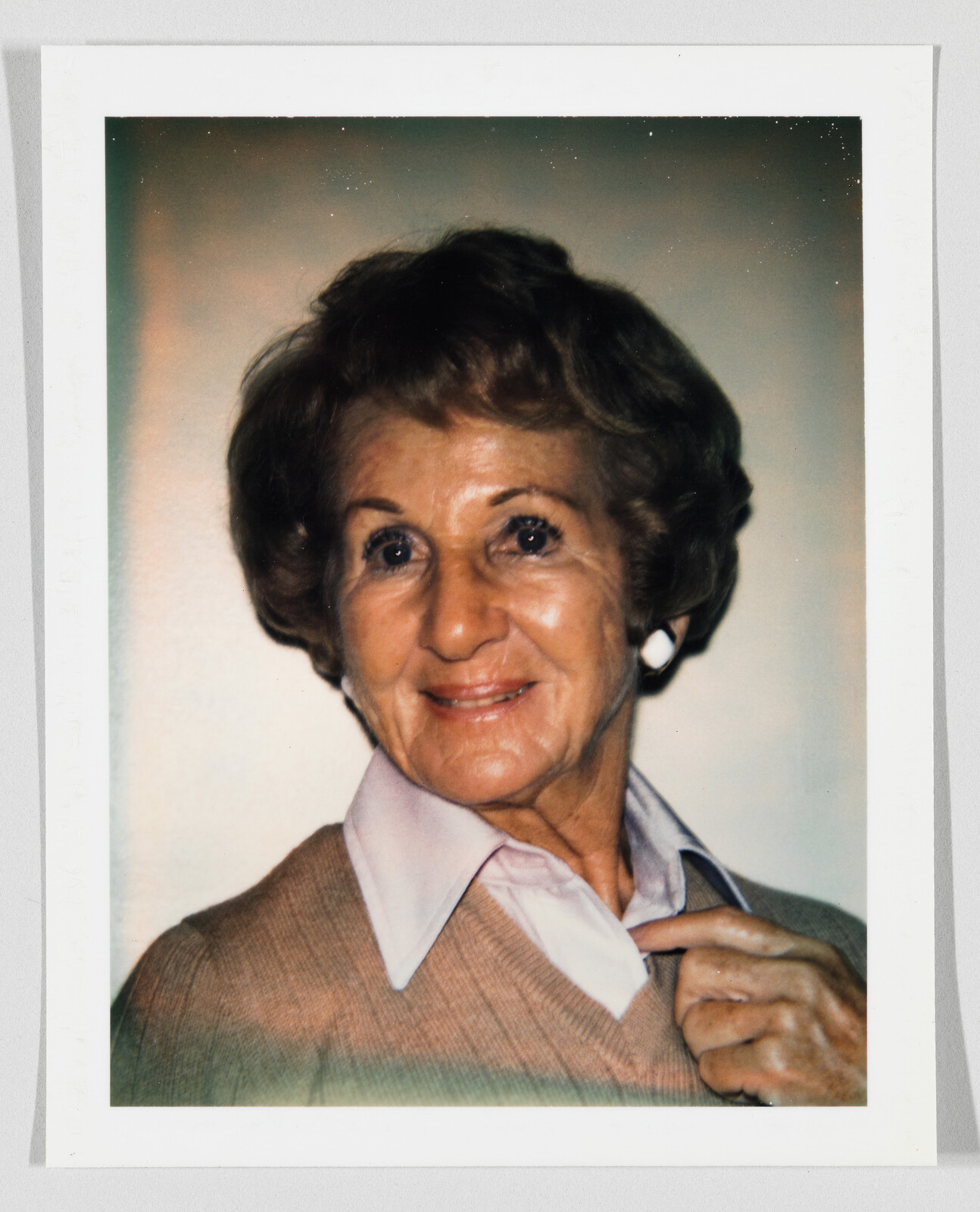 An elderly woman smiles while adjusting her shirt collar and wearing white earrings.