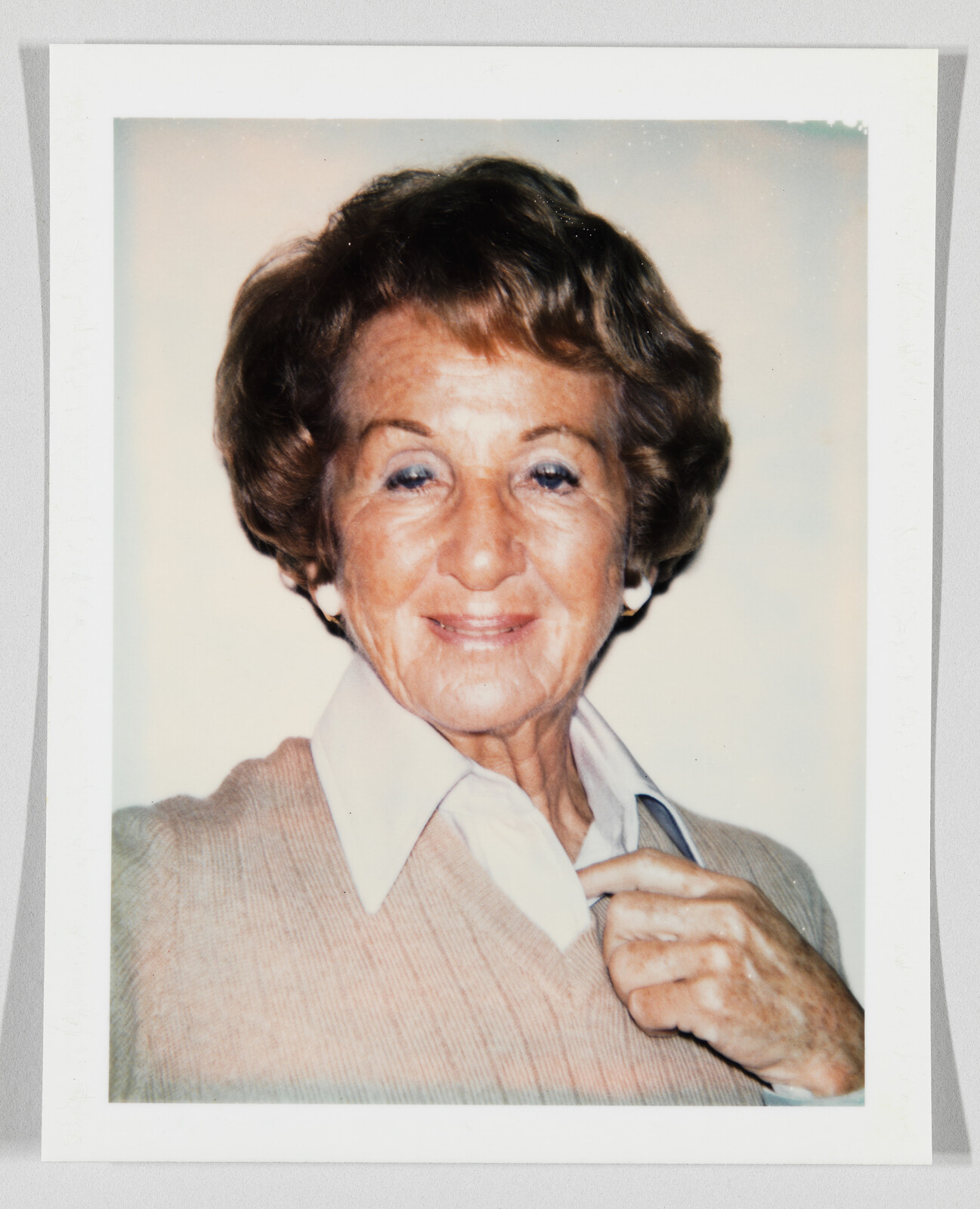 Older woman with short brown hair smiles, wearing a light sweater and collared shirt, holding her collar.