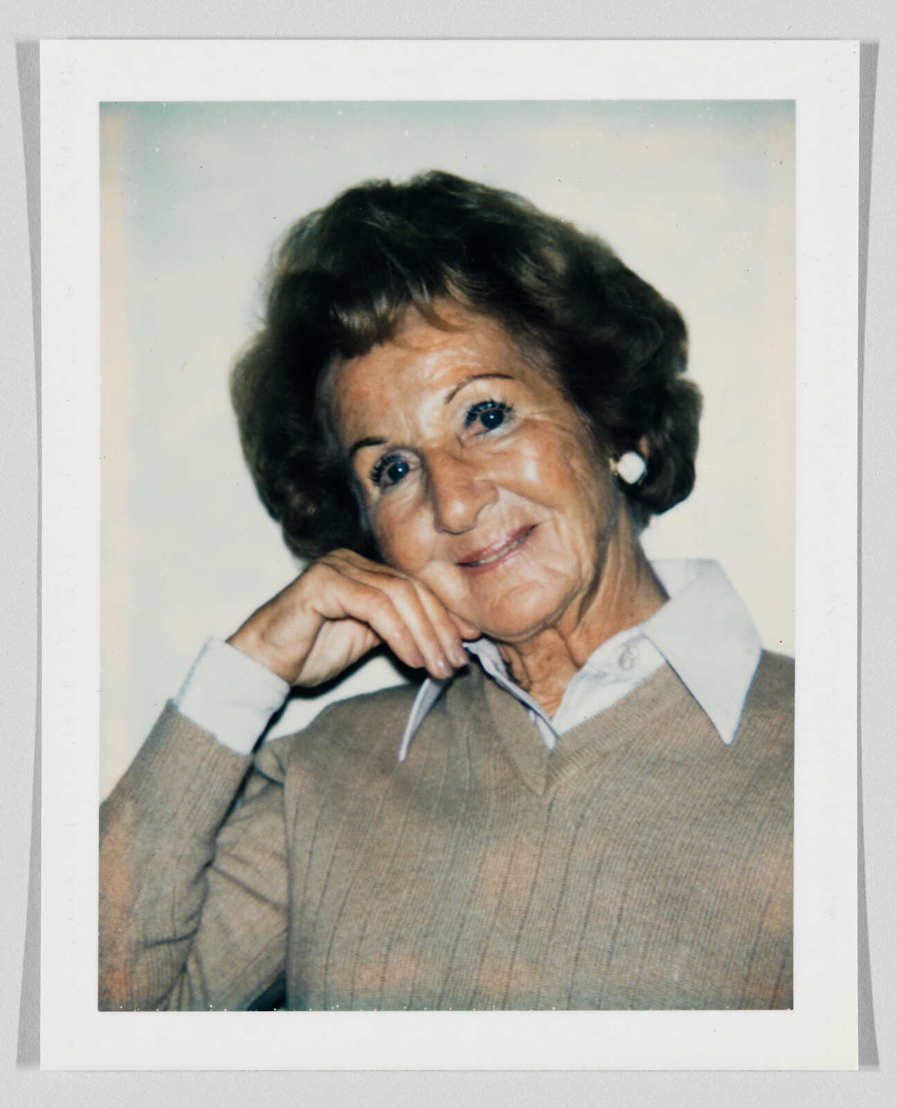 Older woman with short brown hair smiles, resting her chin on her hand, wearing a beige sweater.