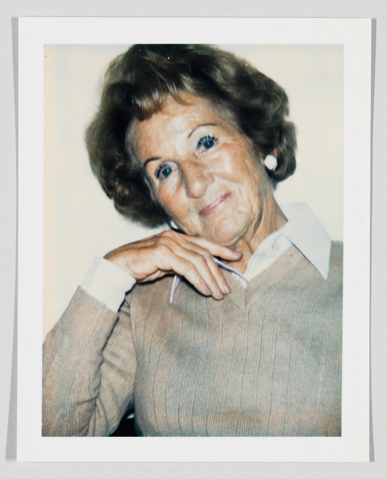 Elderly woman smiling gently with hand resting on her chin wearing a sweater and earrings.