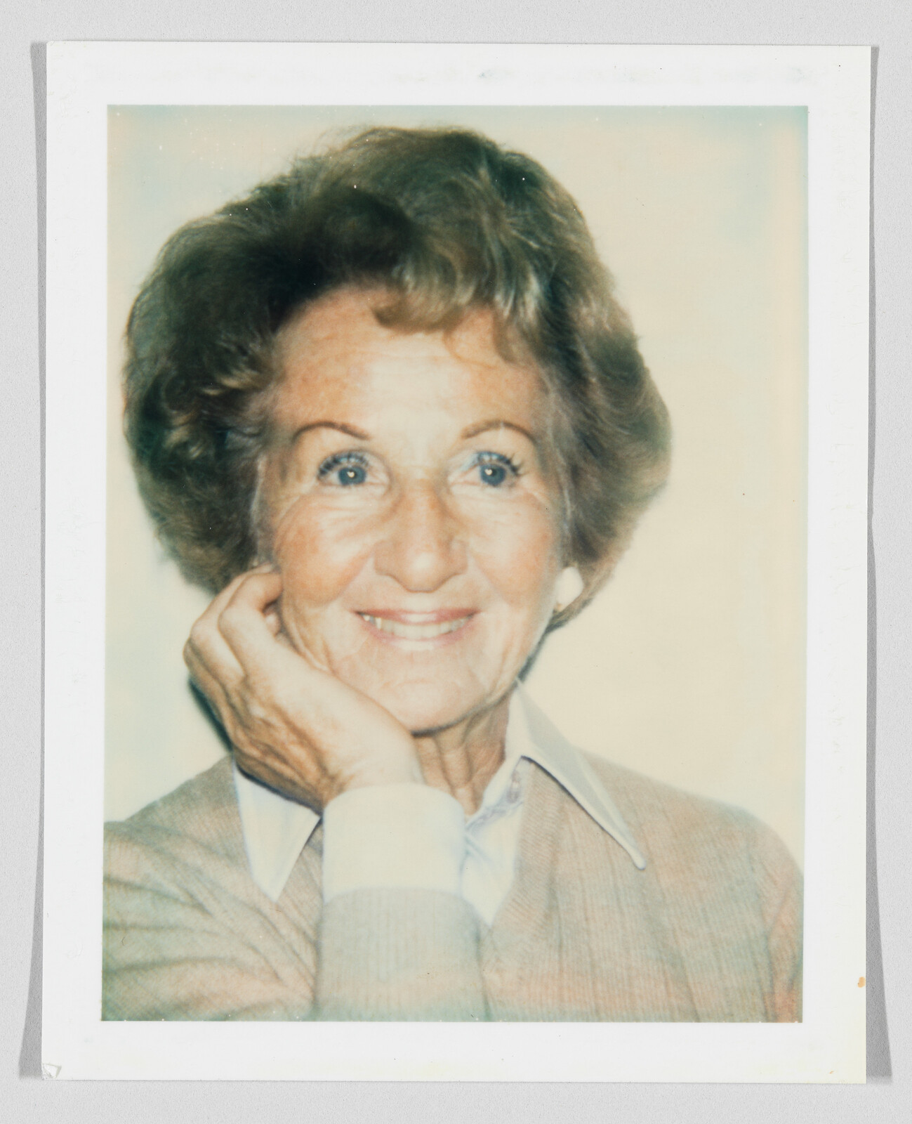 An older woman smiles warmly with her hand resting against her cheek.