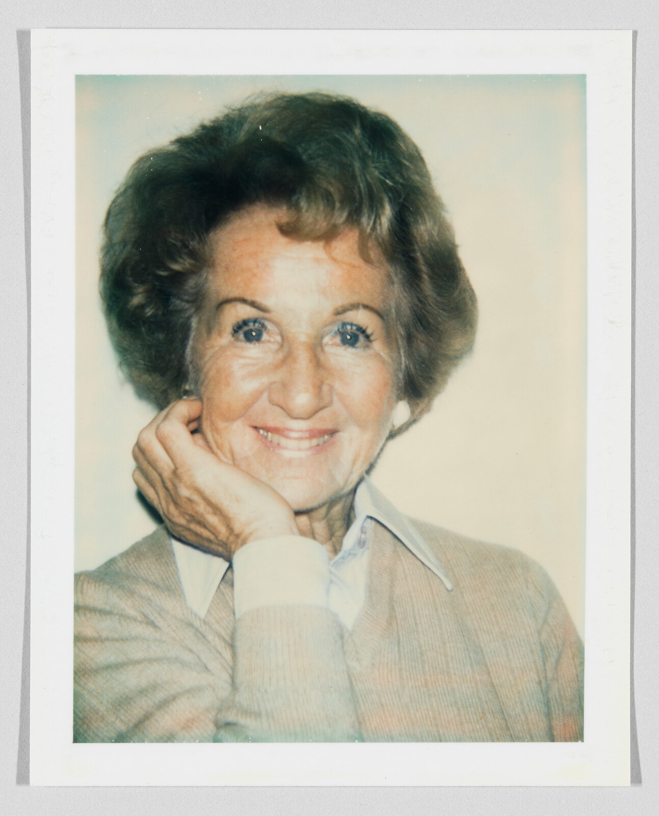 An elderly woman smiles warmly with her hand resting against her cheek.