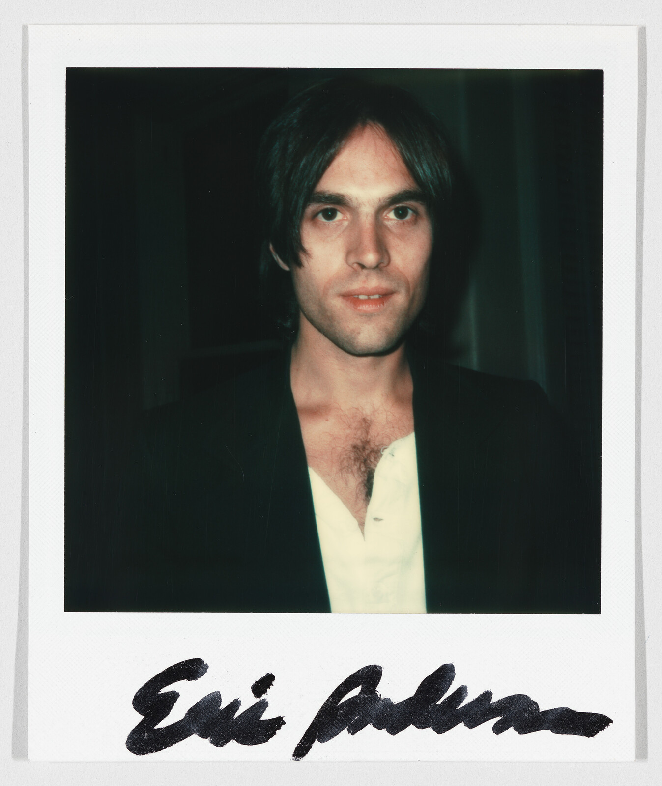 Young man with dark hair looks at camera wearing open white shirt and dark jacket, signature below.