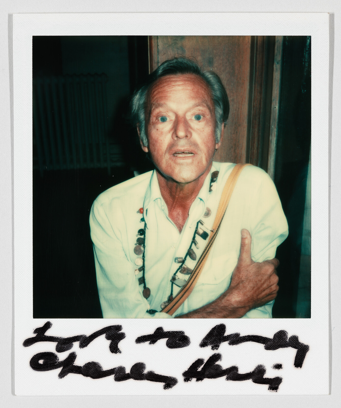 Older man wearing a beaded necklace and strap looks surprised while clutching his arm.