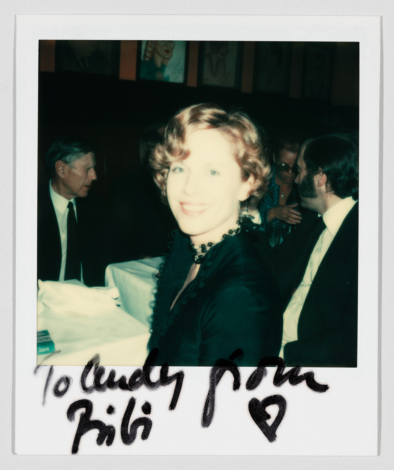 Woman smiling at camera while sitting at a restaurant table with people nearby.