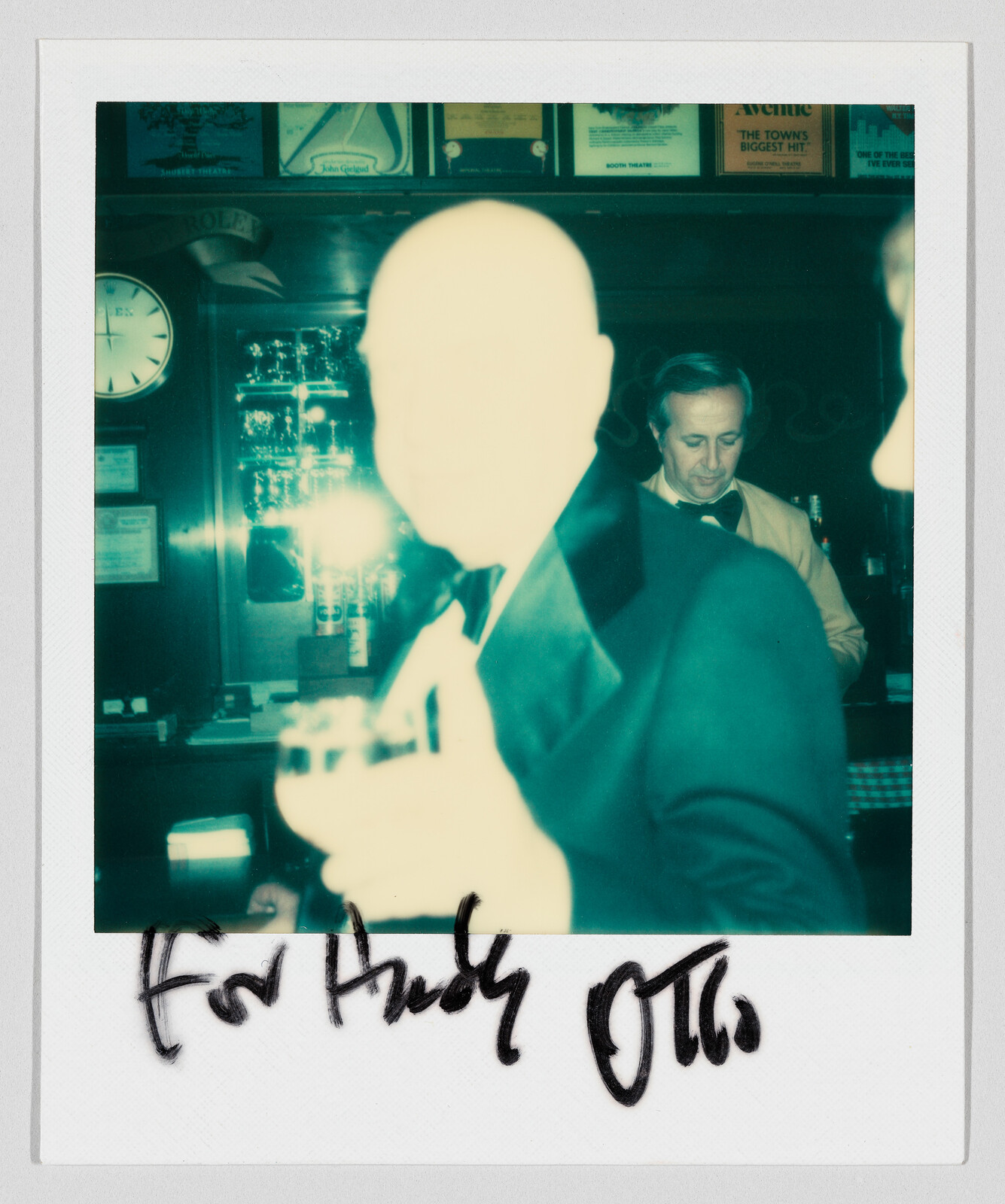 A man in a tuxedo holds a drink at a bar with a handwritten dedication below.
