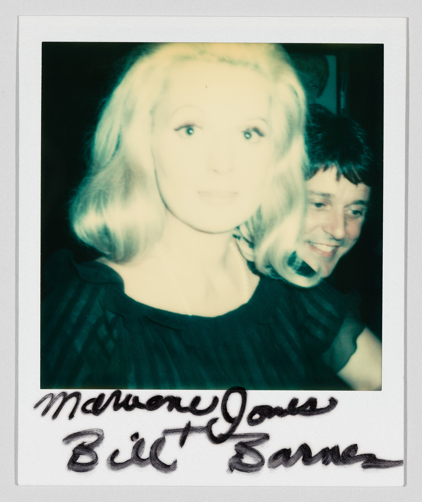 Blonde woman in a dark dress smiling with a man peeking from behind in a Polaroid.