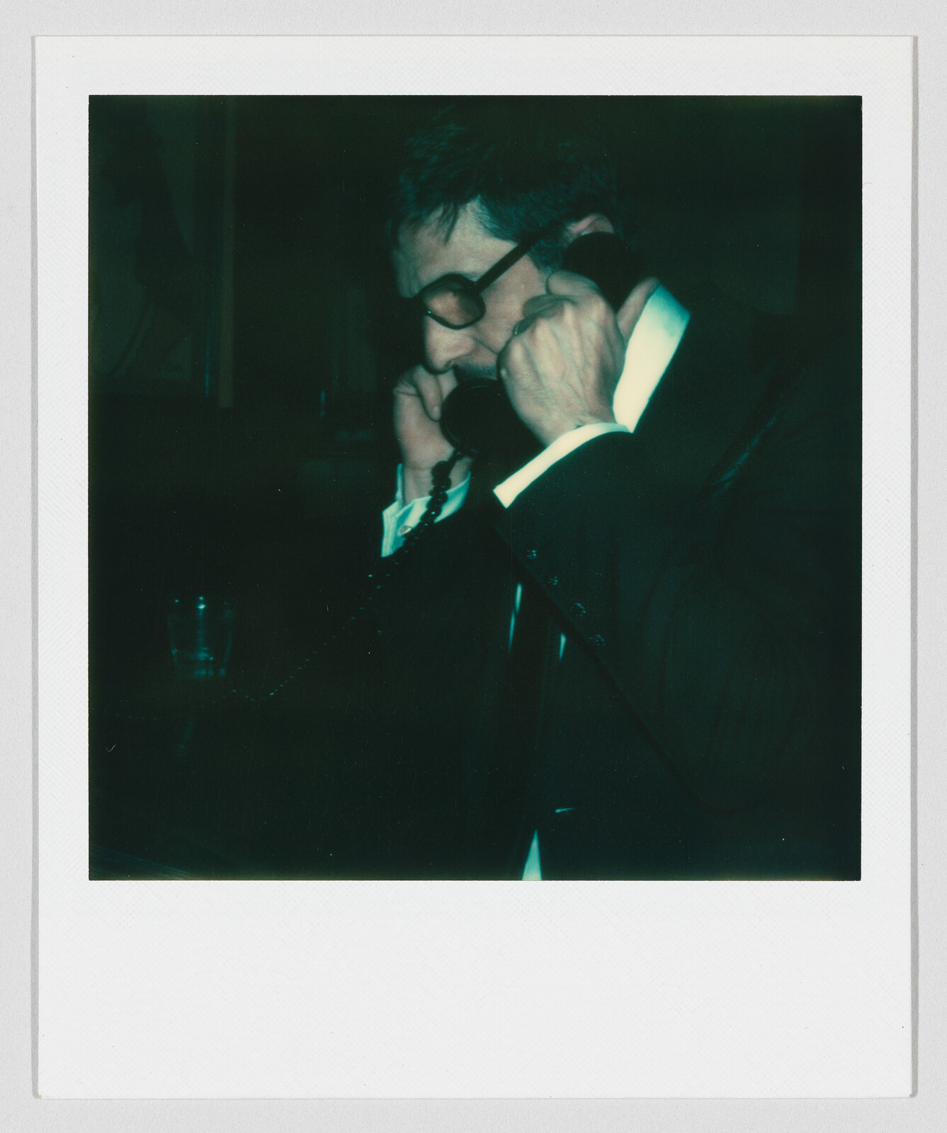A man in a suit and glasses holds a corded telephone receiver to his ear.
