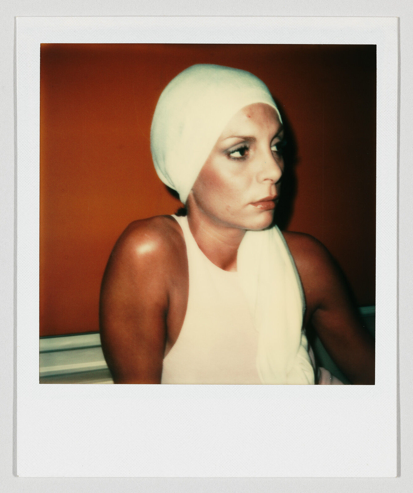 Woman wearing a white headscarf looks to the right with a serious expression against an orange background.