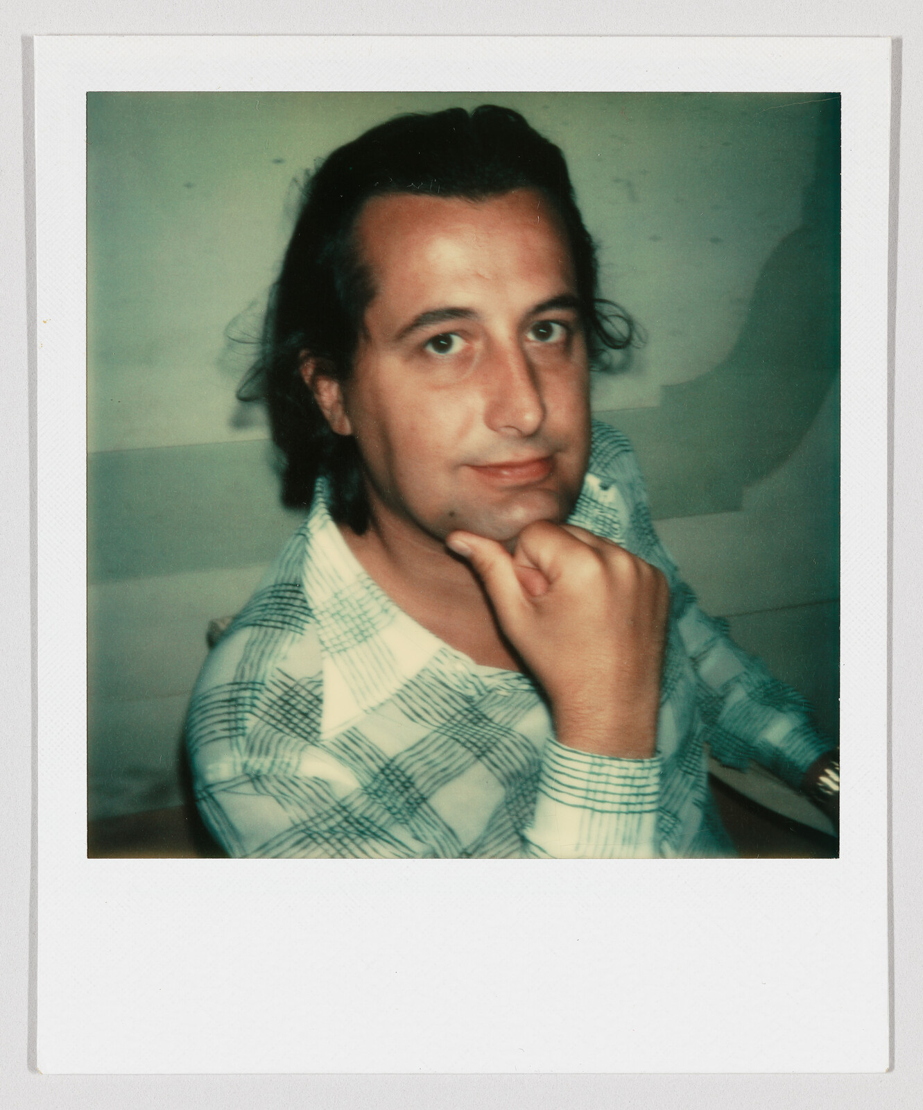 A man wearing a green plaid shirt rests his chin on his hand and looks at the camera.