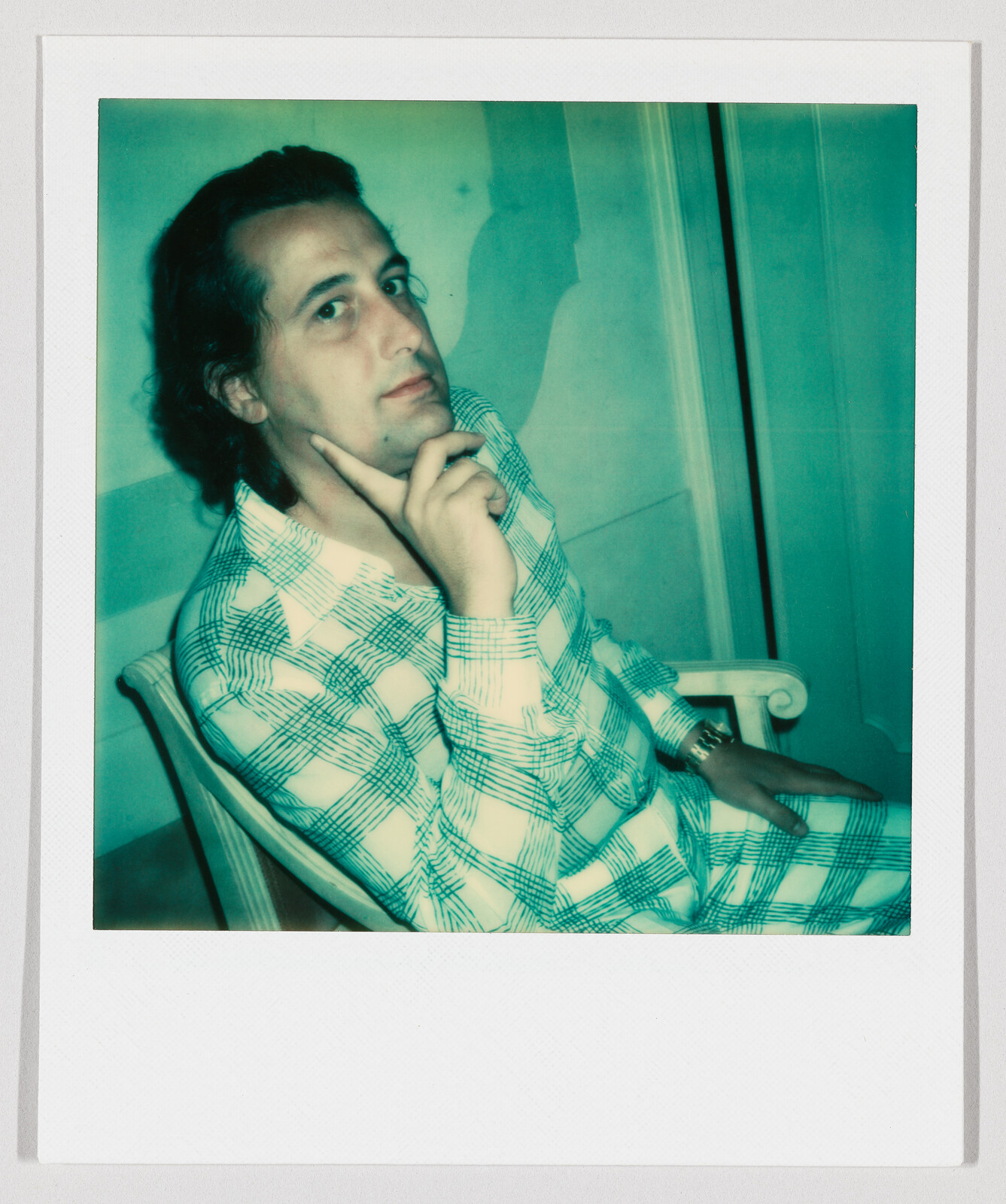 Man in checked shirt sits in a chair, resting chin on fingers and looking directly at the camera.