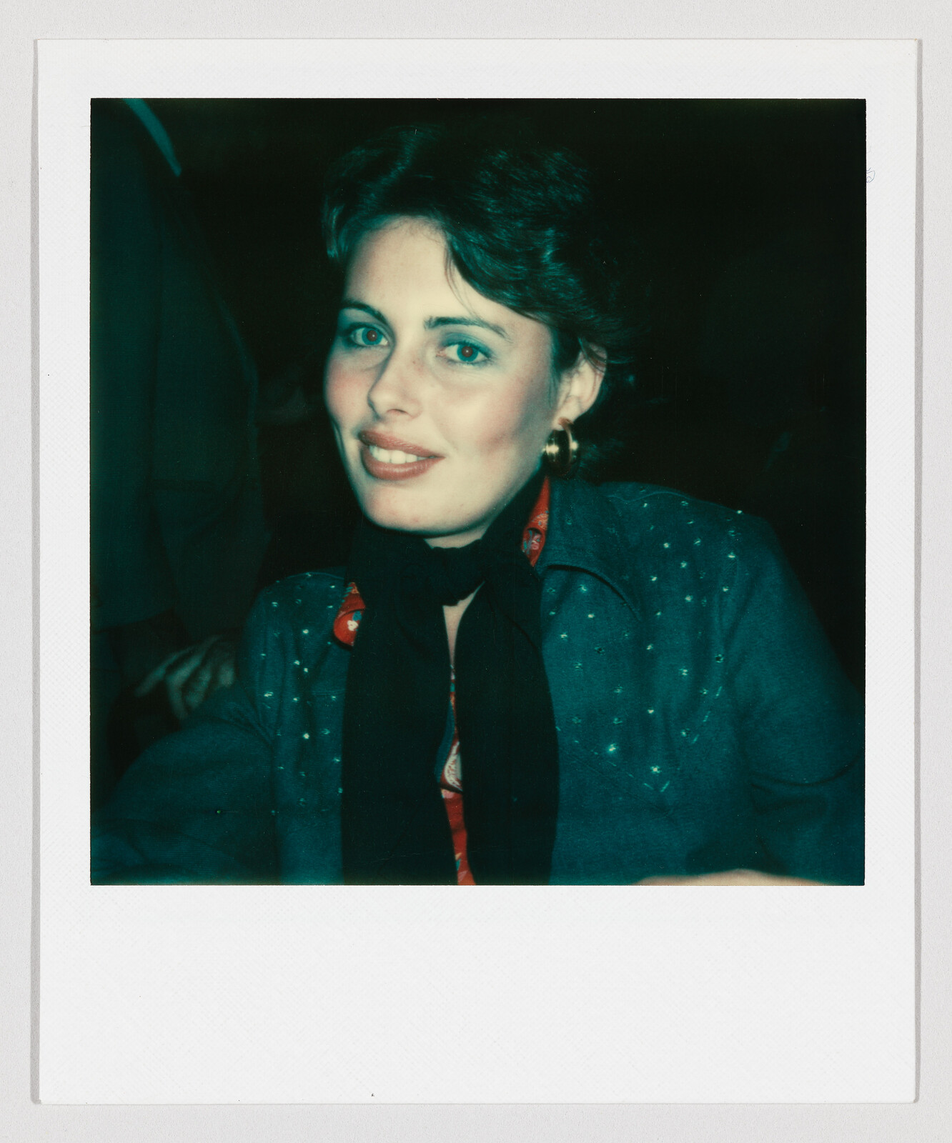 A smiling woman wearing a teal jacket, black scarf, and gold hoop earrings looks toward the camera.