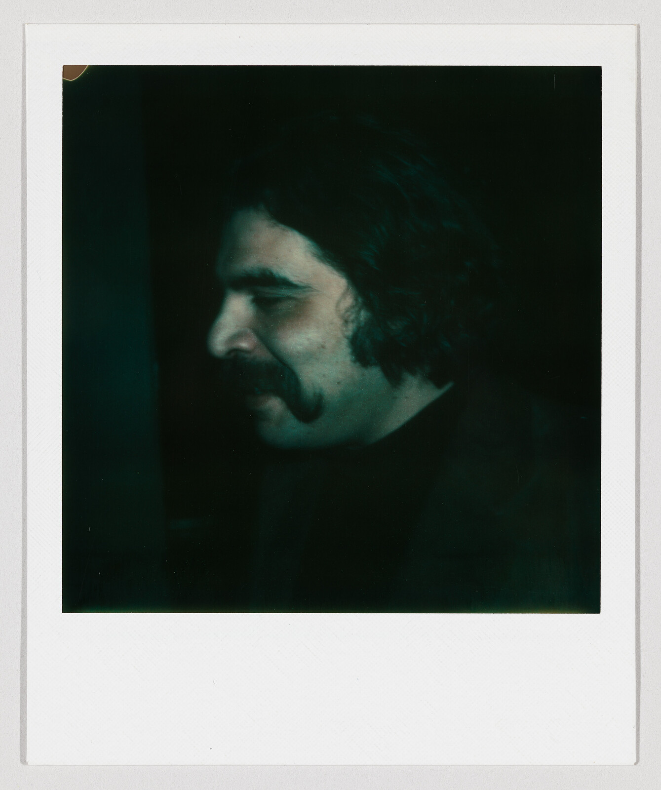 Man with a thick mustache and wavy hair shown in dark side-profile within a Polaroid frame.