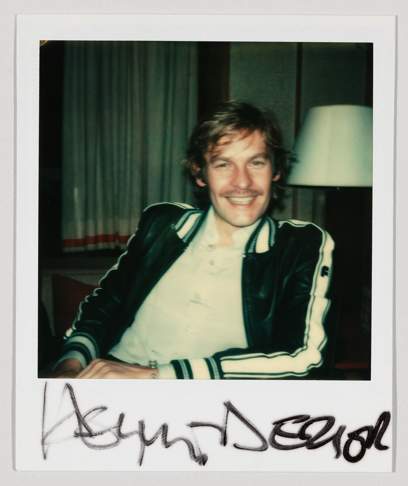 A smiling man in a striped-sleeve leather jacket sits relaxed on a chair indoors.
