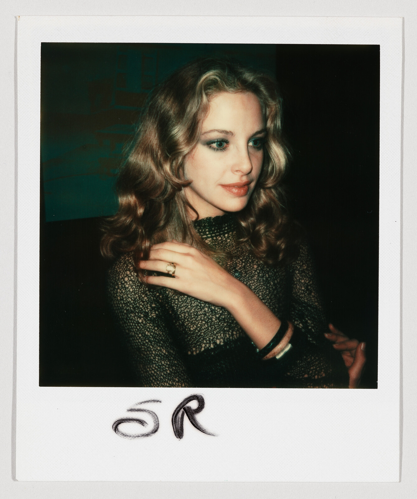 A woman with wavy hair and a black top looks to the side, wearing a ring and bracelets.