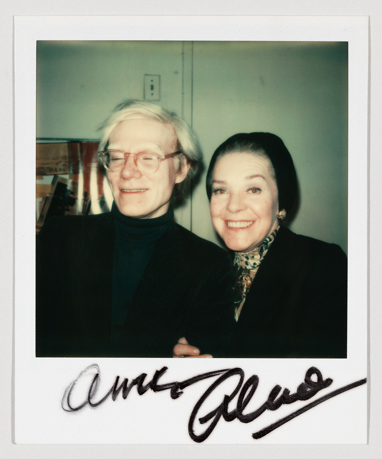 Two people smiling closely together, one wearing glasses and the other wearing a headscarf and scarf.