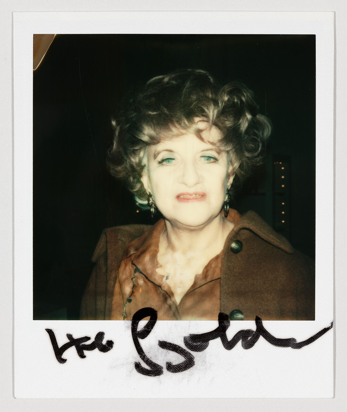 Elderly woman with curled hair and bright makeup wearing a brown coat in a signed Polaroid.