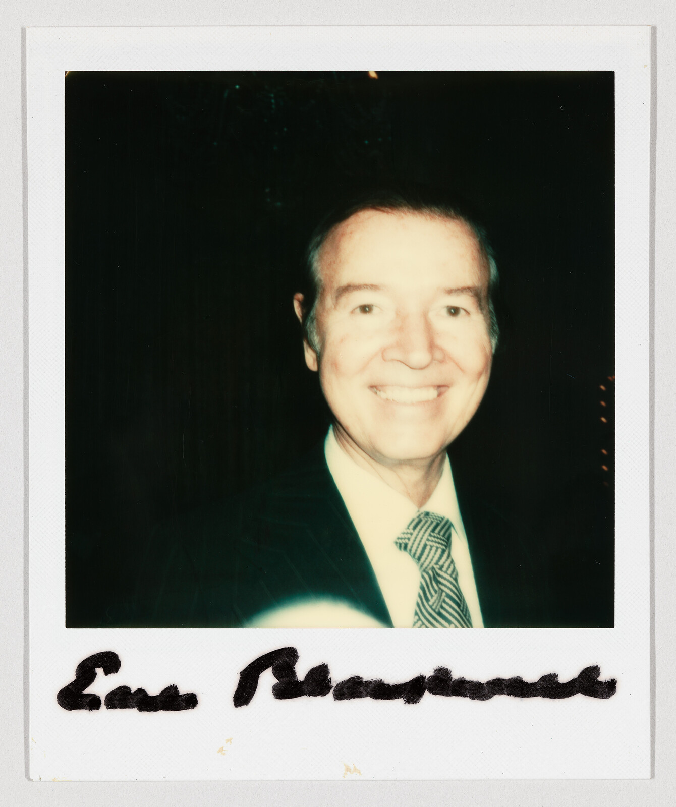 Smiling older man in a suit and patterned tie in a Polaroid with a handwritten signature.