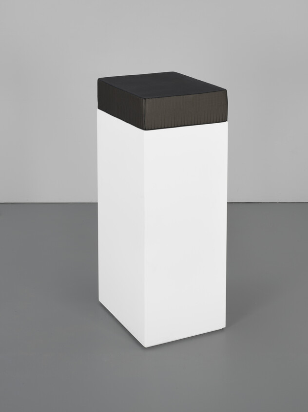 Tall white rectangular pedestal with a black cushioned top standing in a gray room.