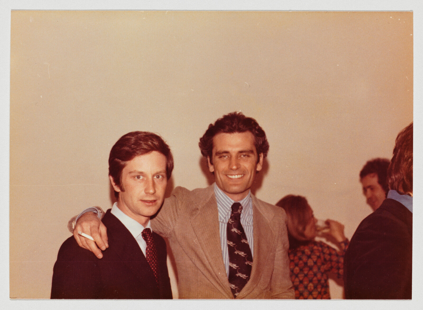 Two men in suits pose together with one man's arm around the other's shoulder and a cigarette.