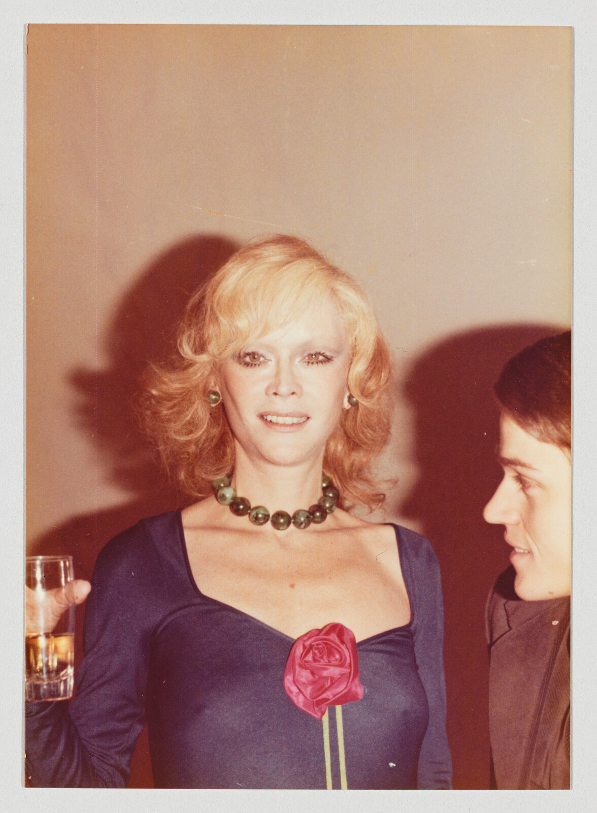 Blonde woman in a blue dress with a red rose pin and green necklace holding a drink.