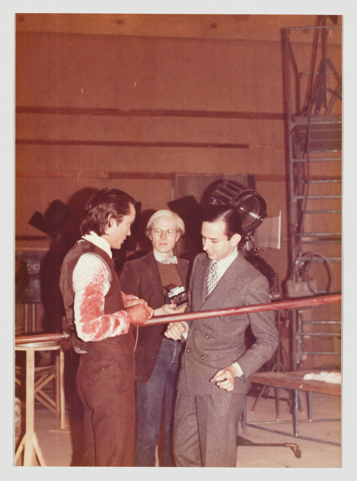 Three men stand on a film set, one in a blood-stained costume holding a prop pole.