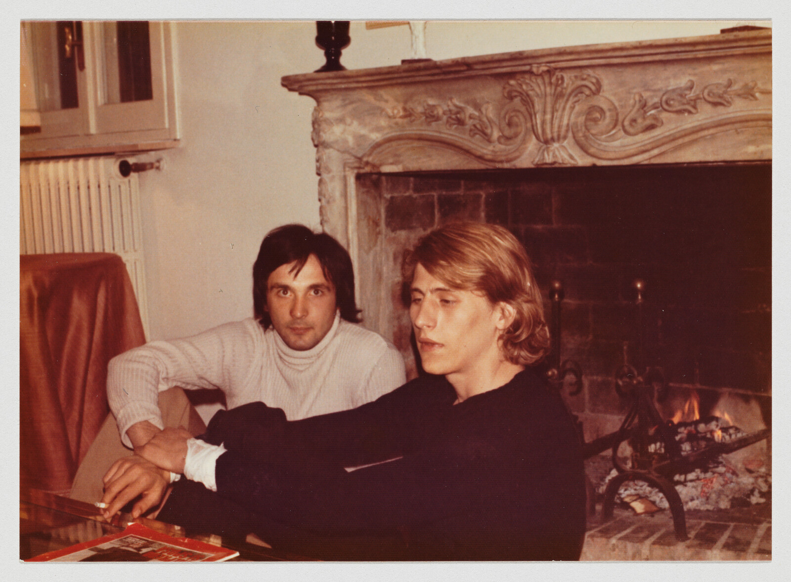 Two people sit in front of a lit fireplace, one wearing a white sweater and the other in black.