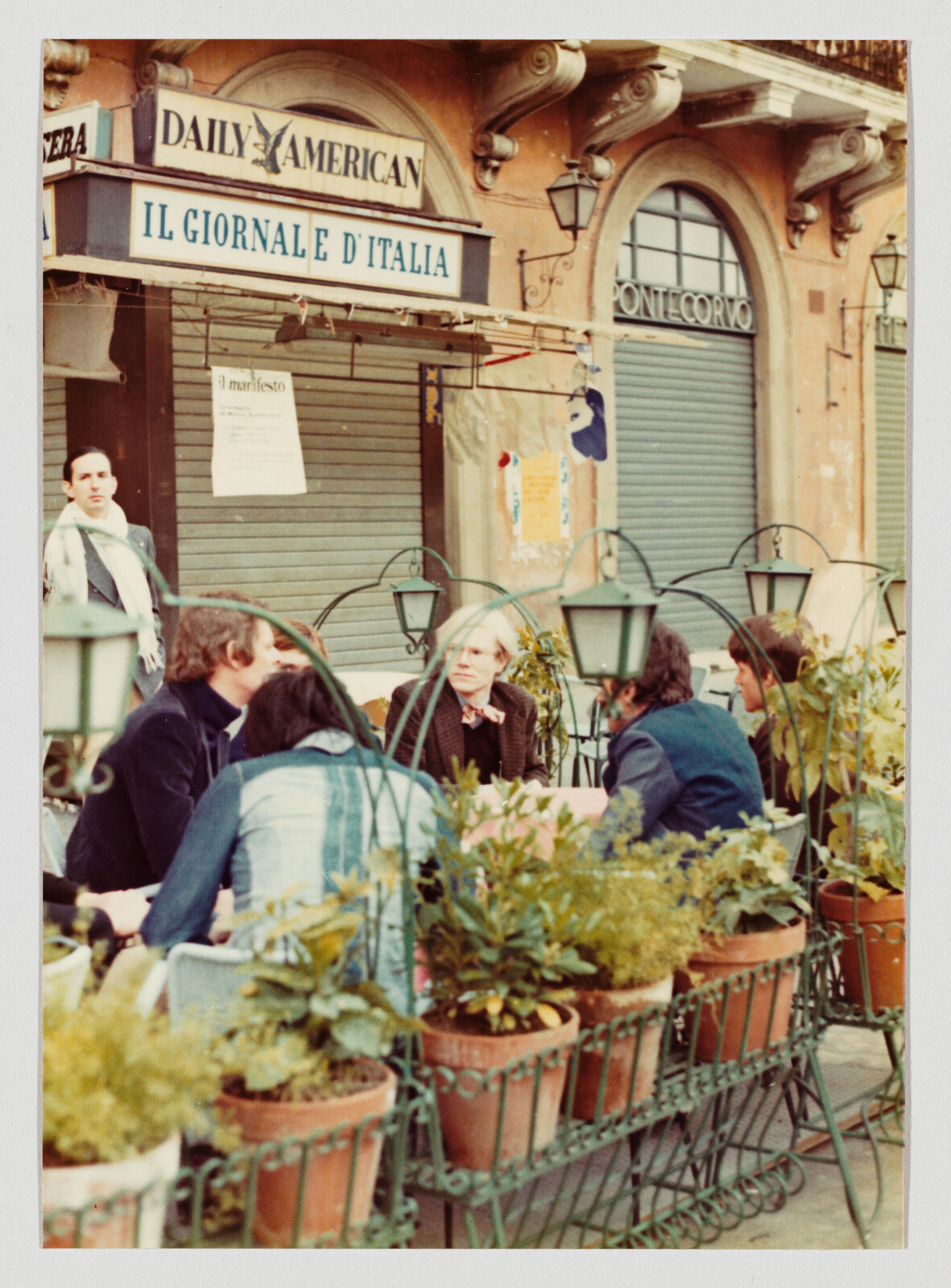 A group of people sit and talk at an outdoor café table behind a row of potted plants.