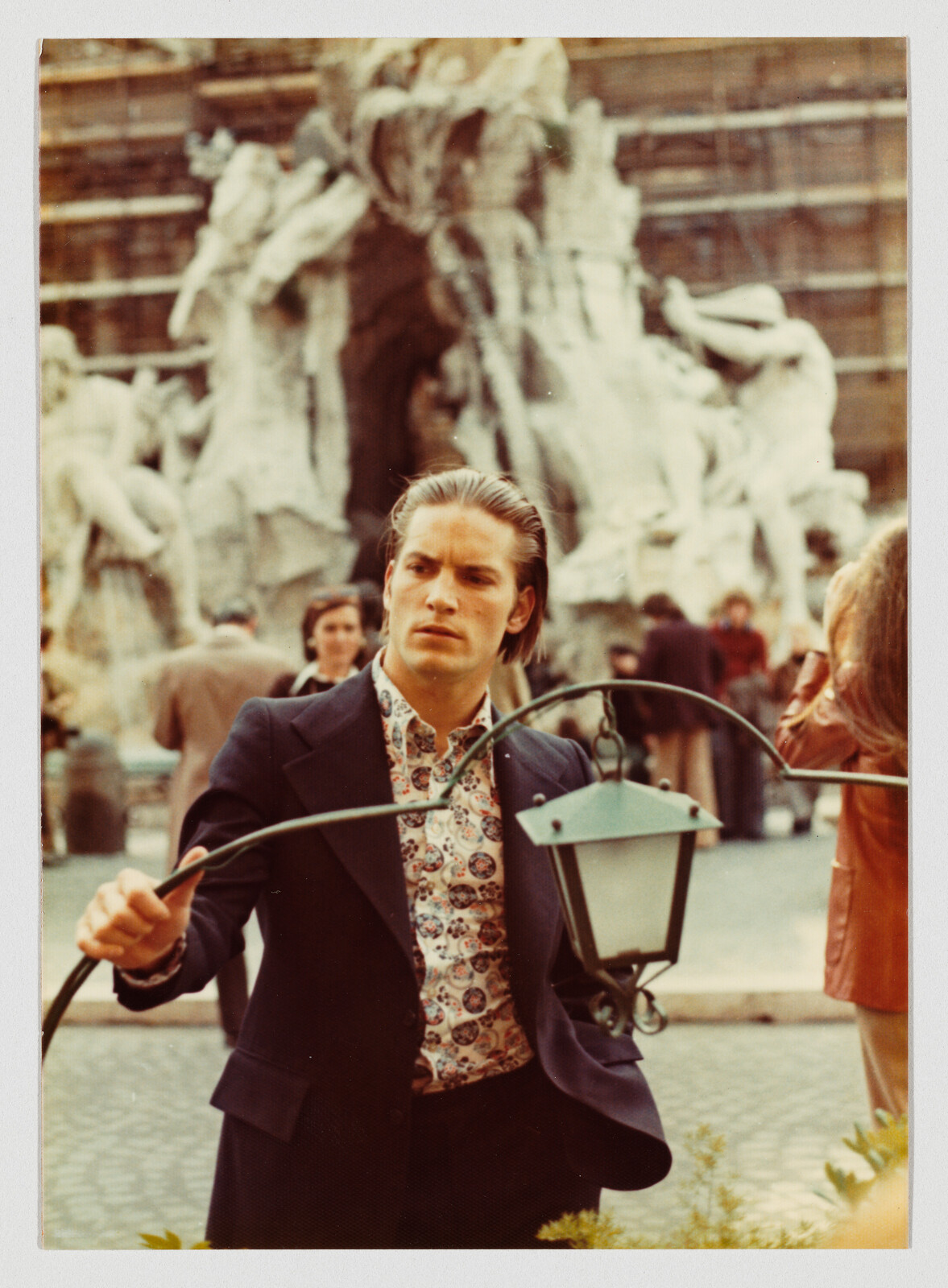 Young man in patterned shirt holds a lamp arm while standing near a large stone fountain.