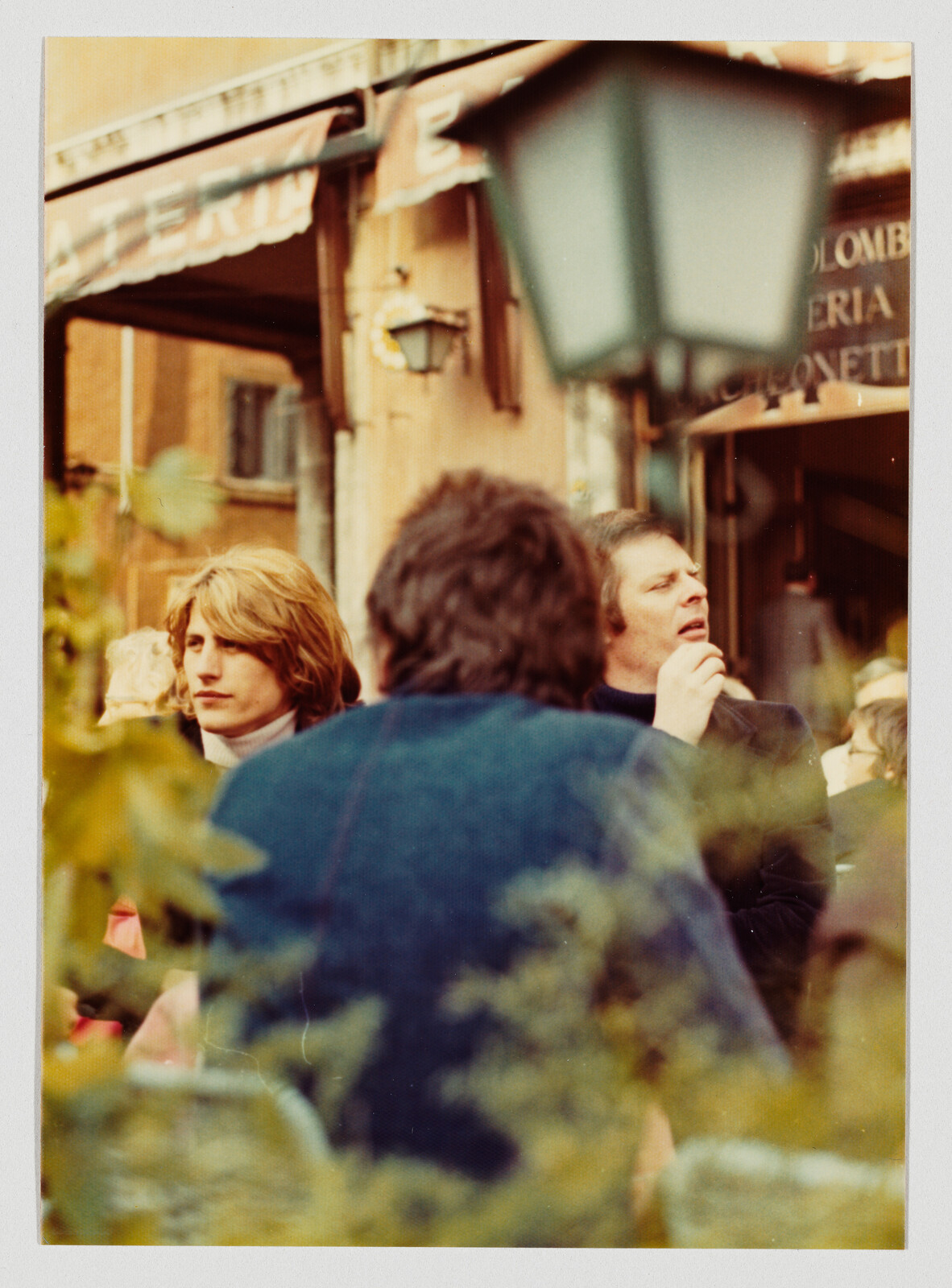 Two men sitting at an outdoor café, one man holding a cigarette near his mouth.