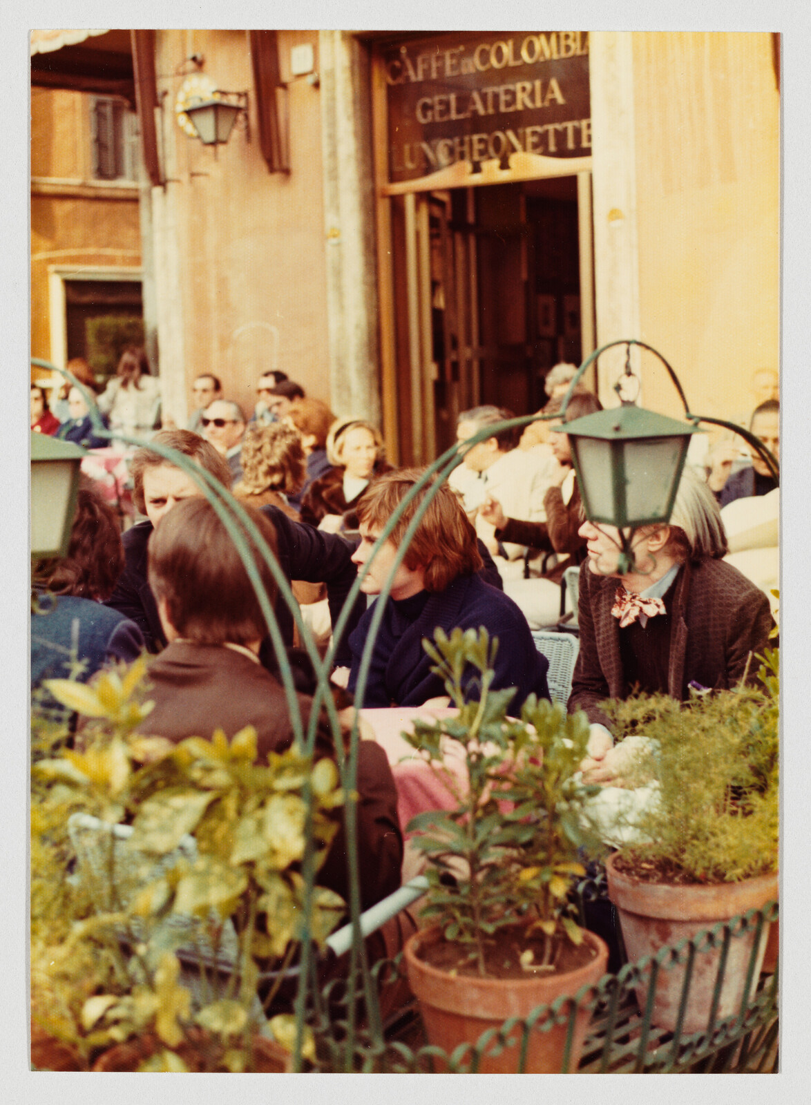 People sit and talk at a busy outdoor café terrace with potted plants in foreground.