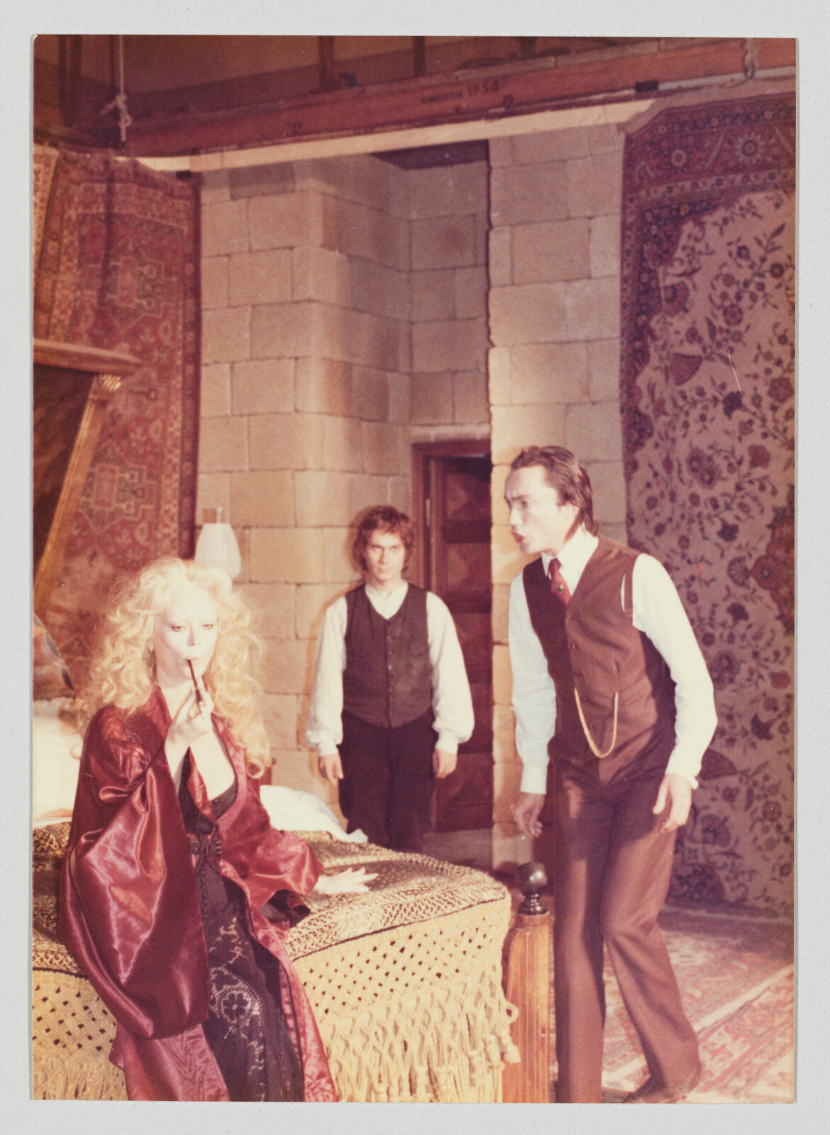 Blonde woman in a silk robe smokes while two men stand and speak in a decorated room.