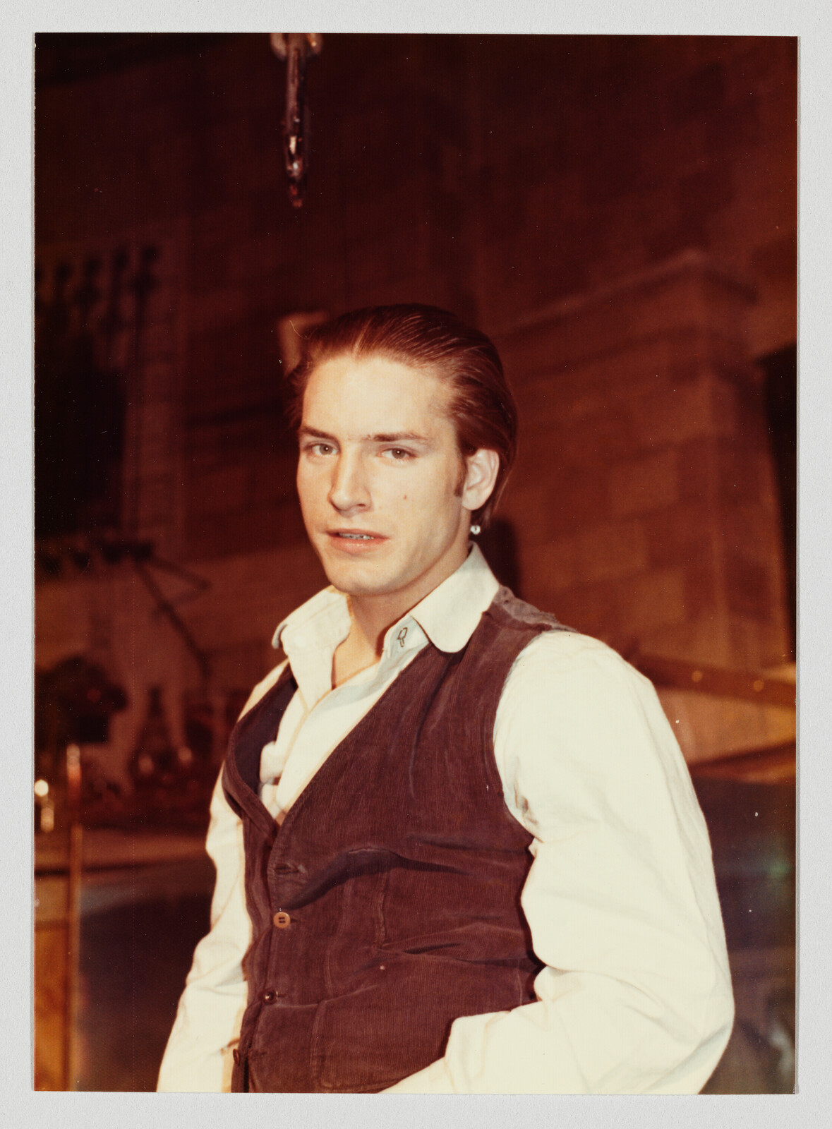 Young man wearing a white shirt and dark vest stands looking directly at the camera indoors.