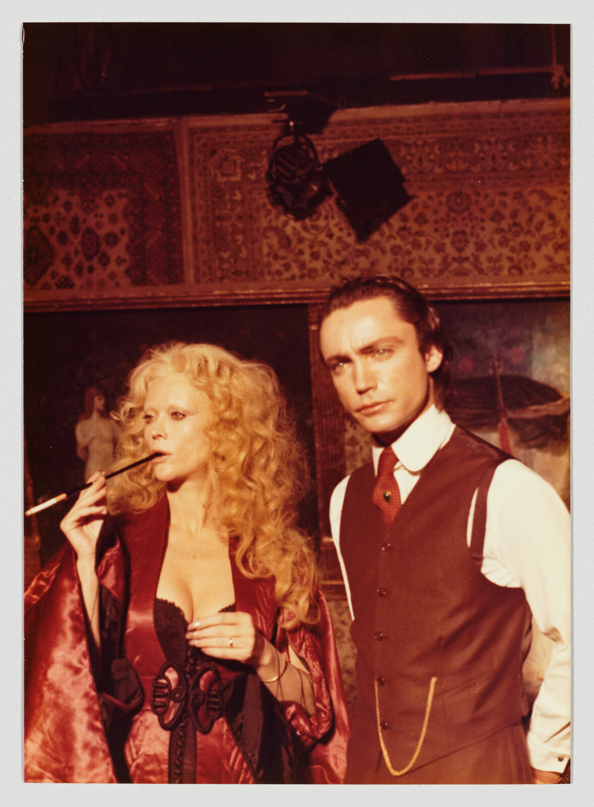 A woman in a silk robe holding a long cigarette holder beside a serious man in vest.
