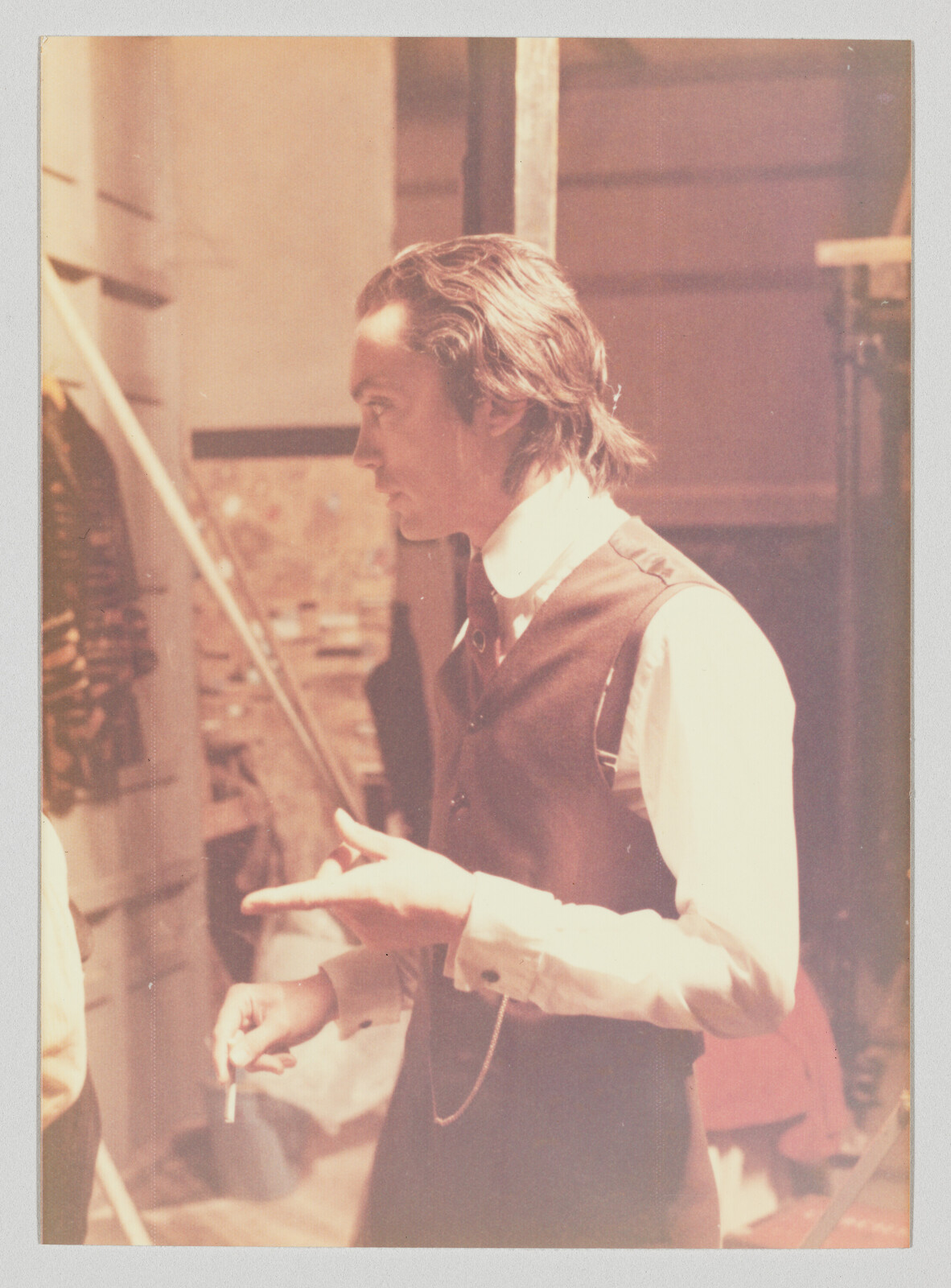 A man in a vest gestures with one hand while holding a cigarette in the other.