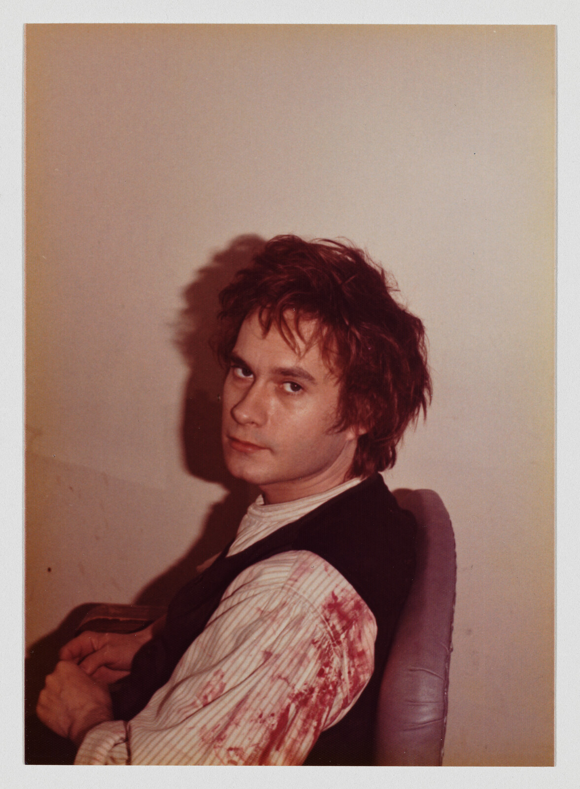 A young man sits in a chair looking back over his shoulder with a bloodstained sleeve.