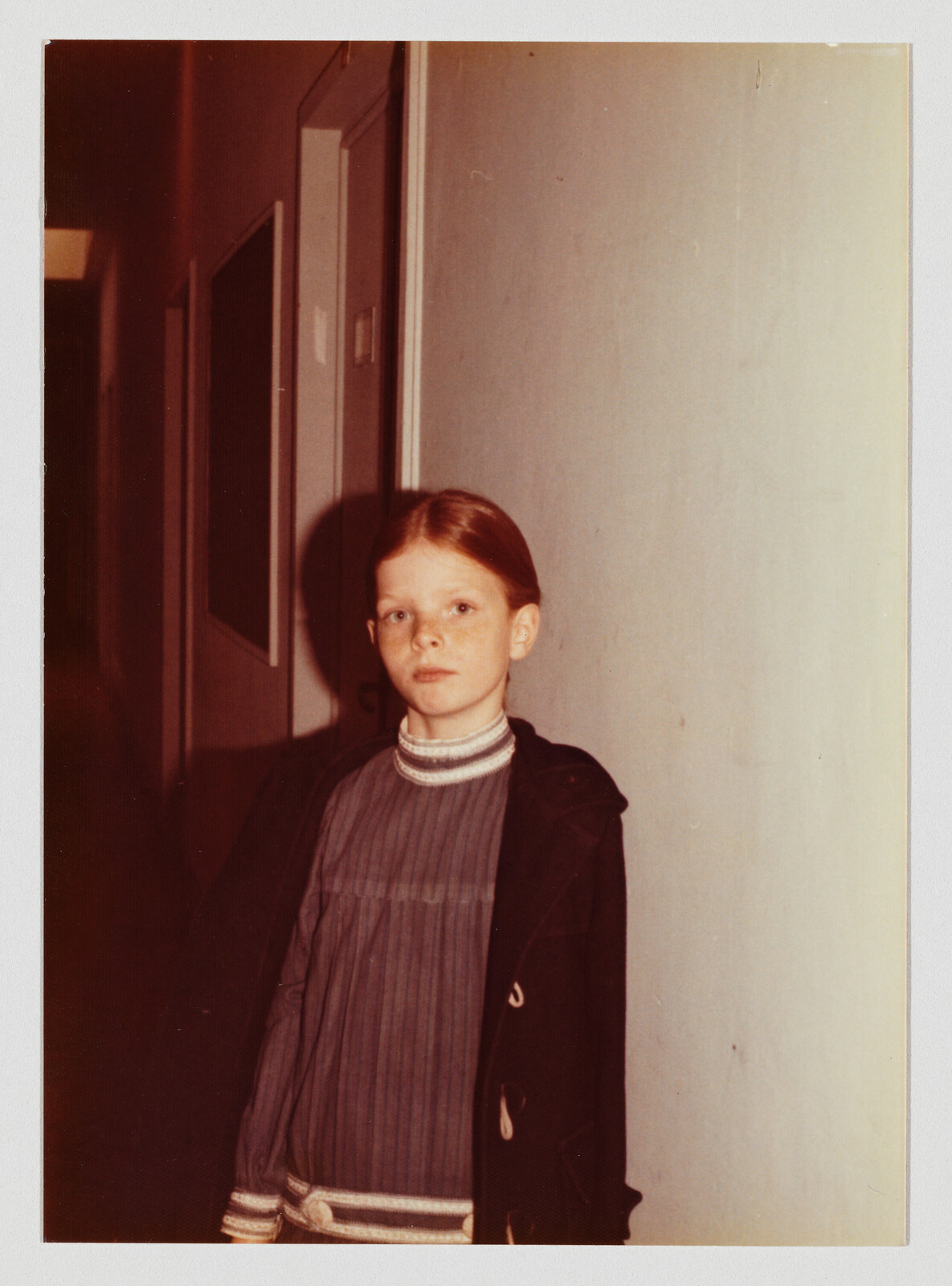 Young girl with red hair stands in a hallway, wearing a striped sweater and dark coat, looking forward.