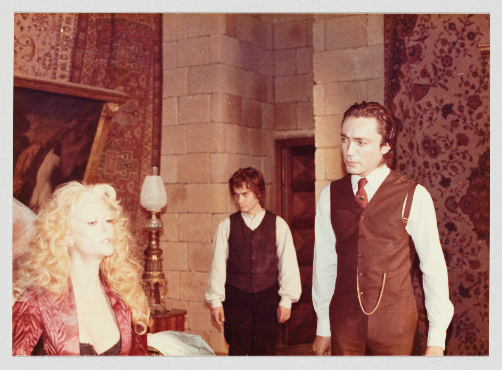 A man in a brown vest confronts a seated blonde woman while another man watches.