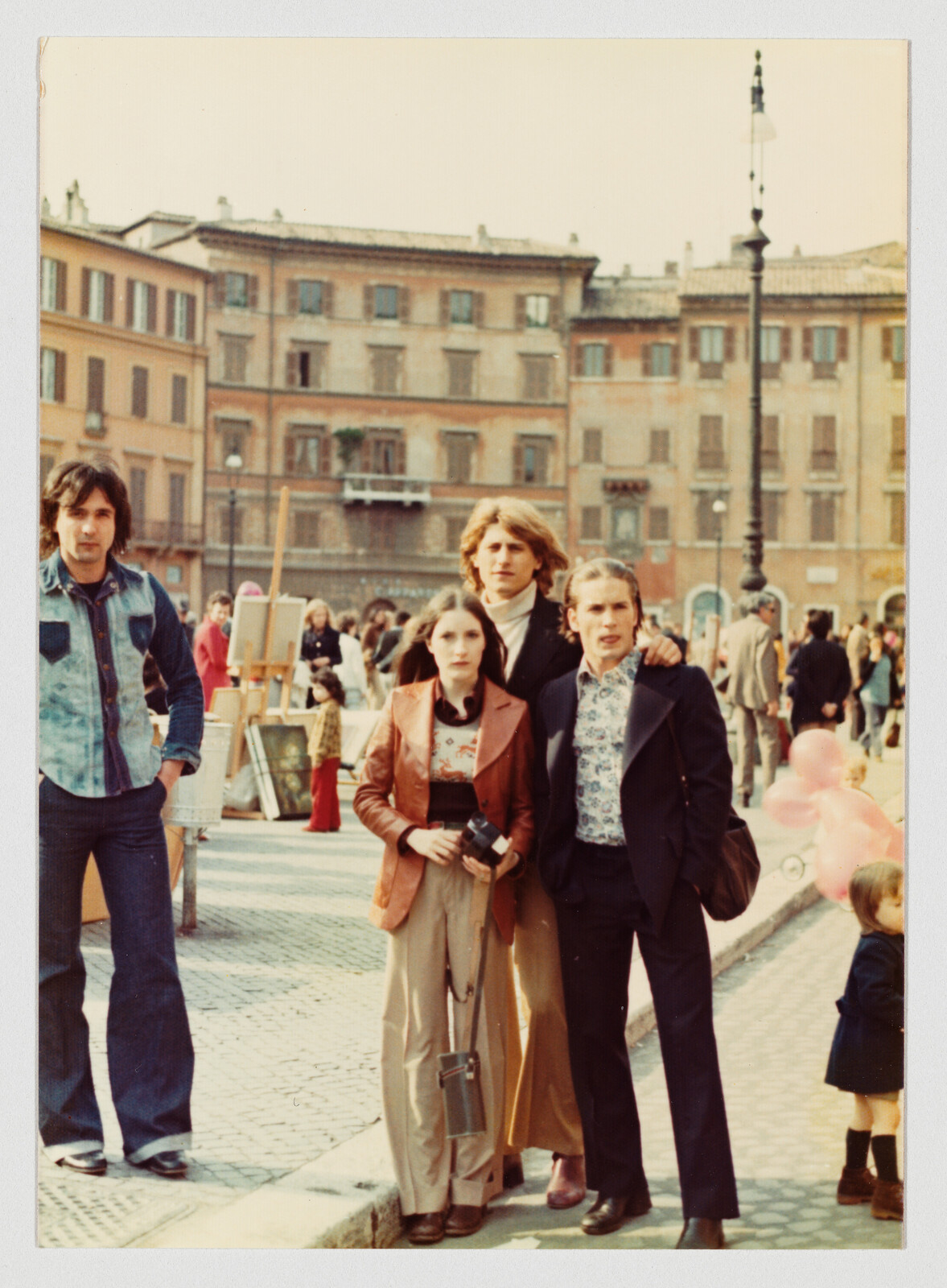 Three adults in 1970s clothing stand together in a busy city square, with people and buildings in the background.