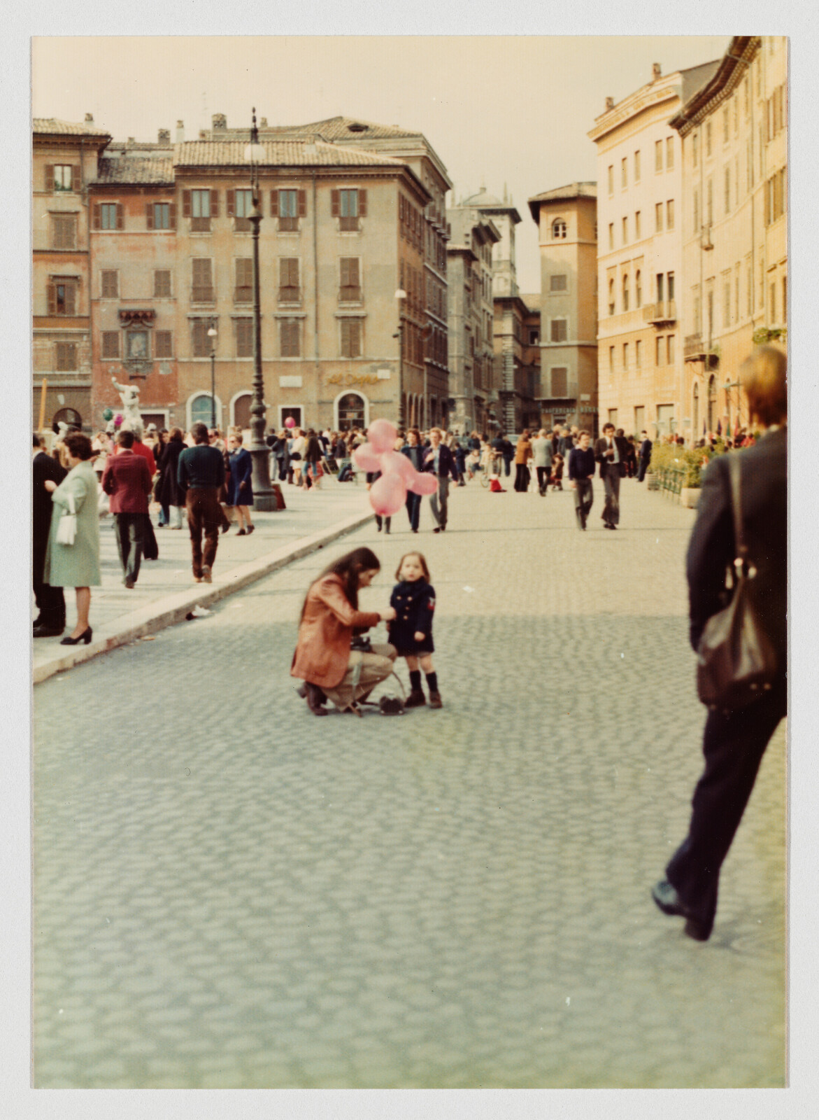 A woman crouches to adjust a toddler's shoes beside a stroller in a busy cobblestone plaza.
