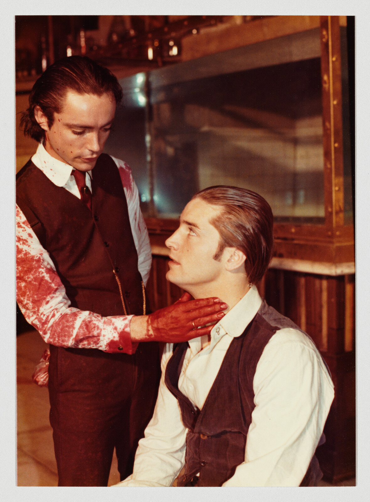 A man with bloodied hands gently holds another man's throat while they stare at each other.