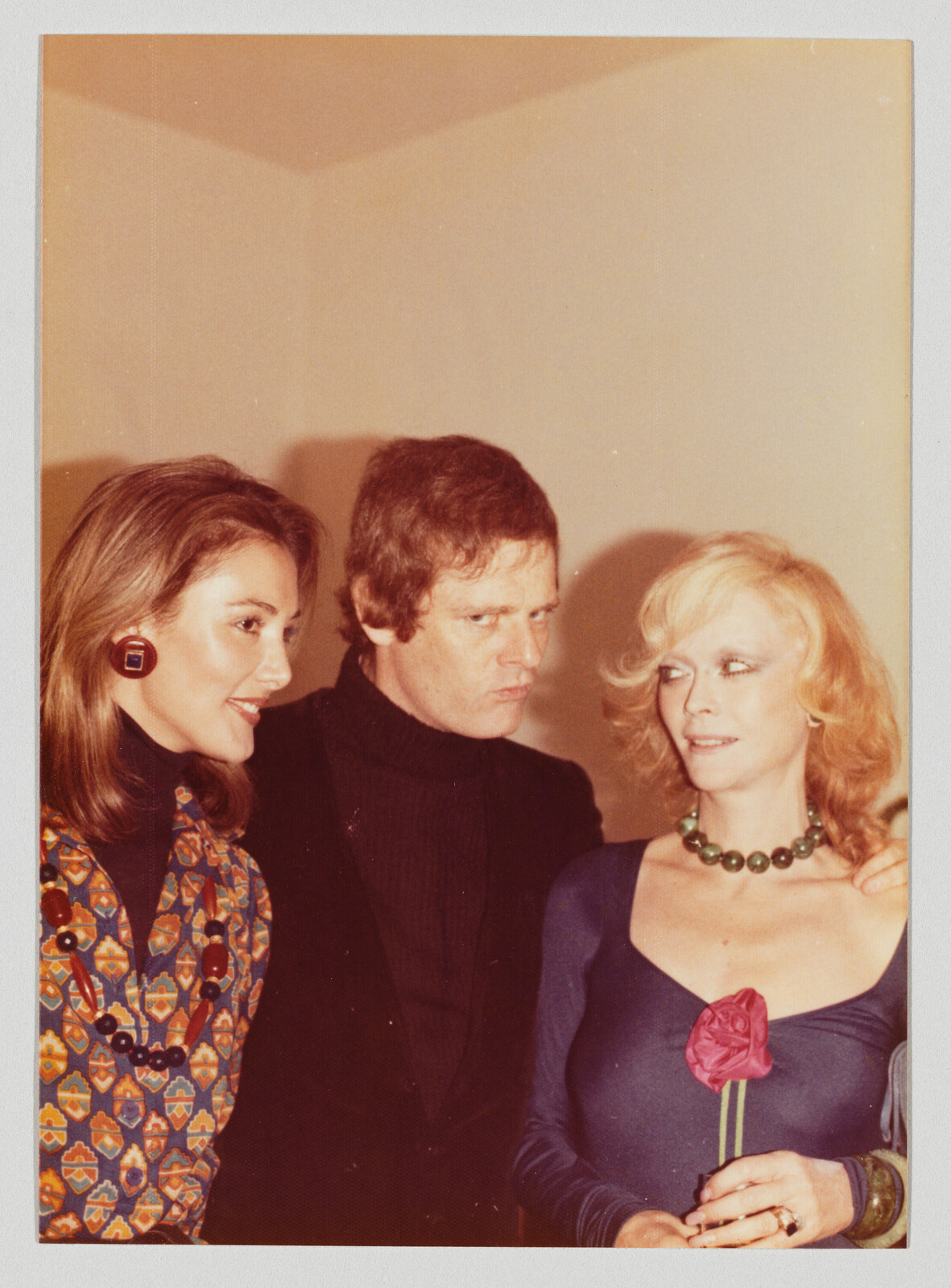 Man looks skeptically at woman holding a pink rose while another woman smiles beside them.
