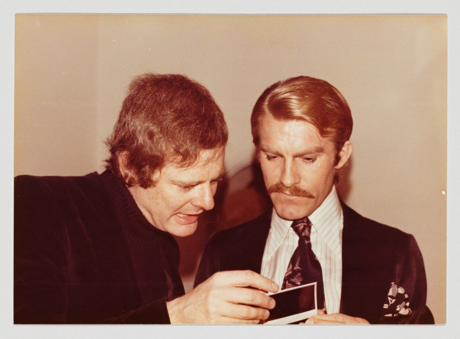 Two men closely examine a small photograph while discussing its contents.