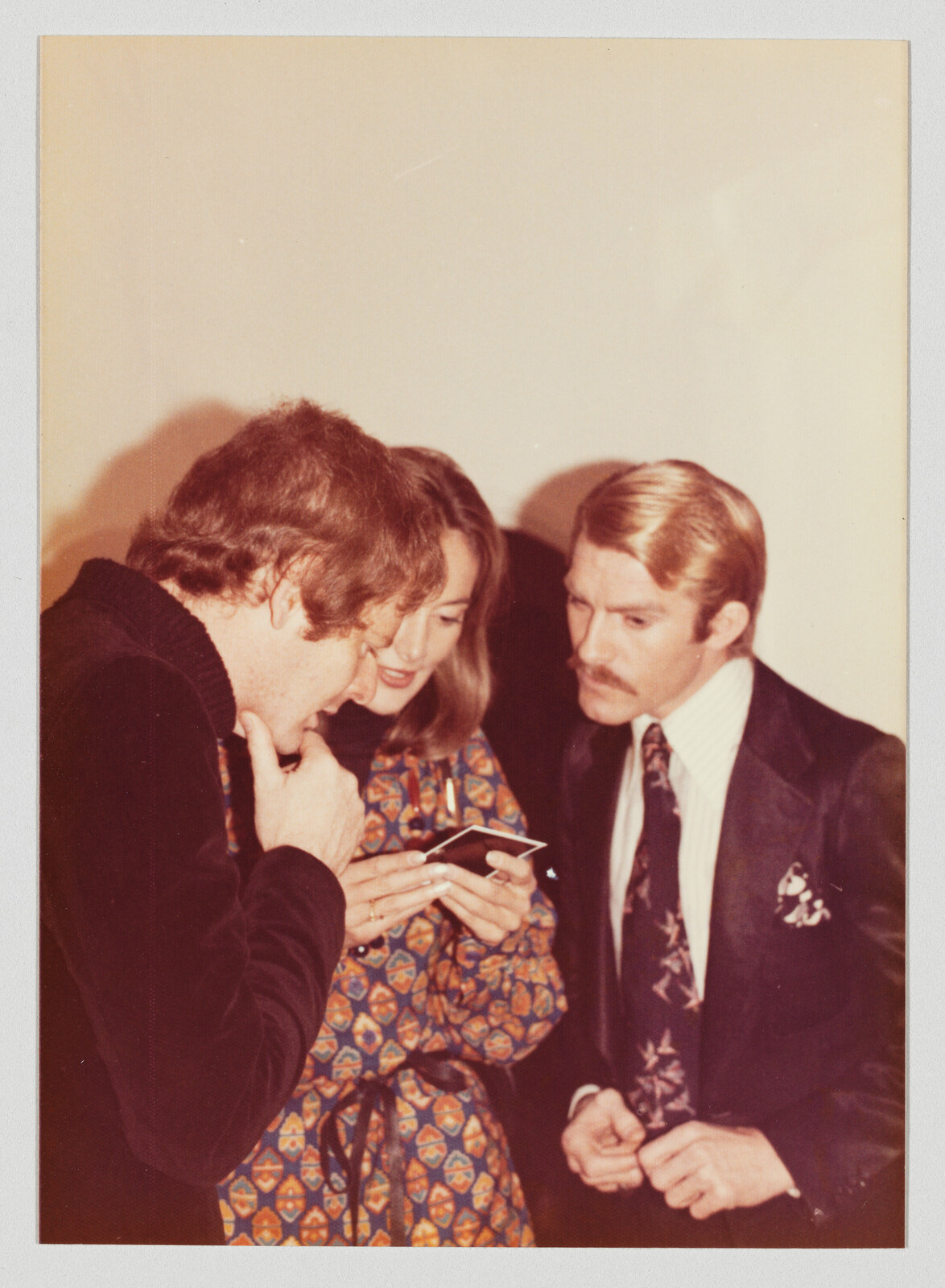 Three people closely examine a small photograph while standing together at an indoor gathering.