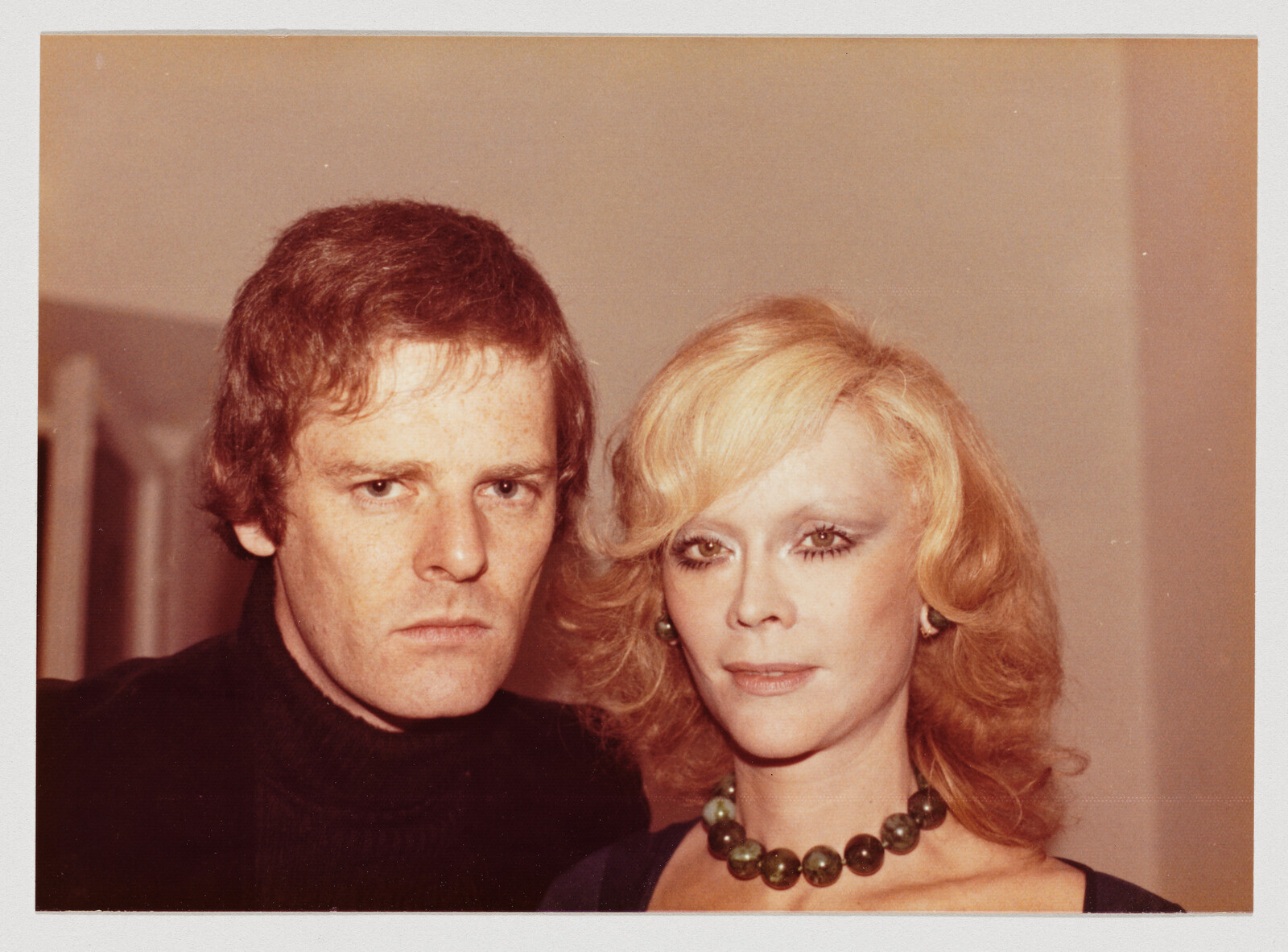 A man and a woman stand close together looking at the camera with neutral expressions.