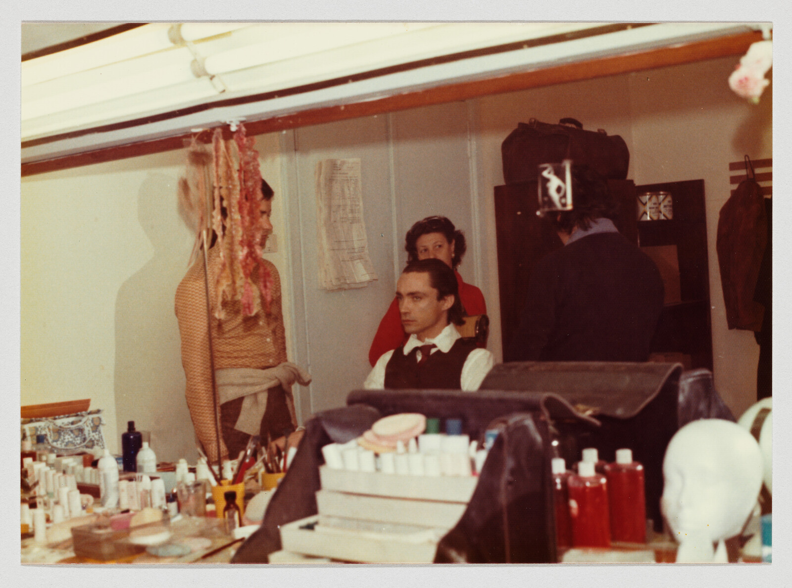 A man in a vest sits in a dressing room while others prepare his hair and makeup.