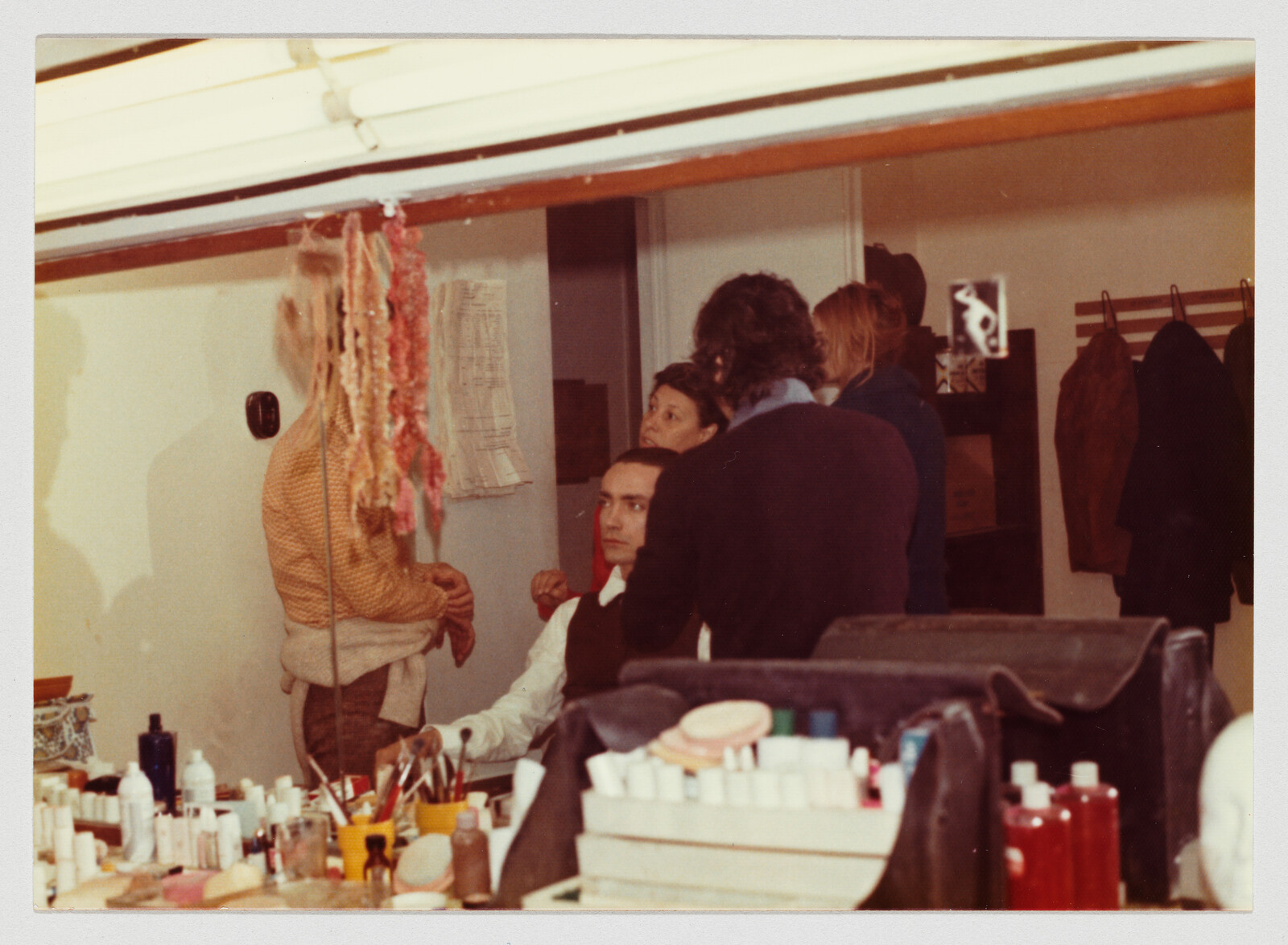 A man sits at a makeup table while several people stand around him in a dressing room.