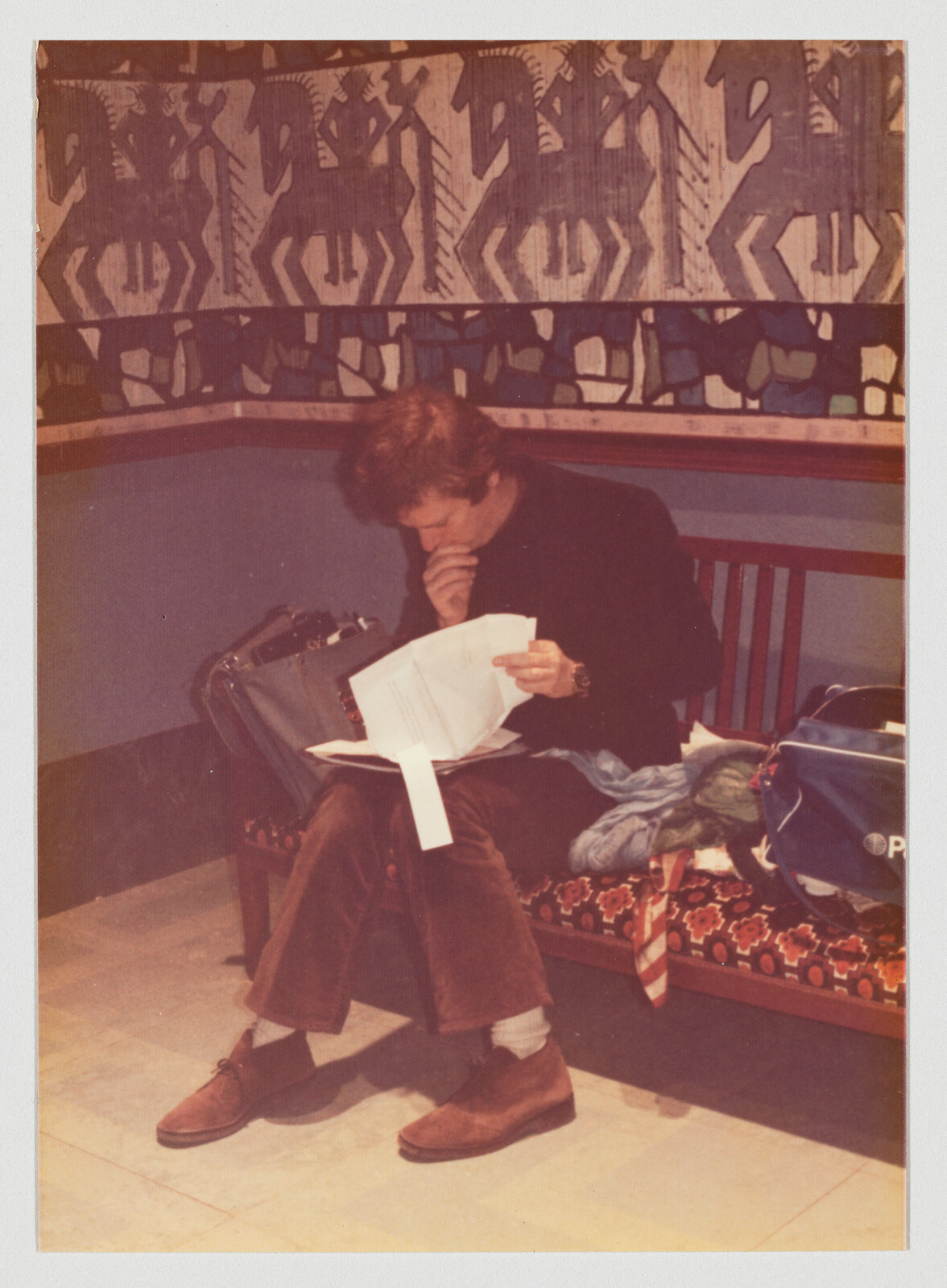 A young man sits on a patterned bench reading documents with a travel bag beside him.