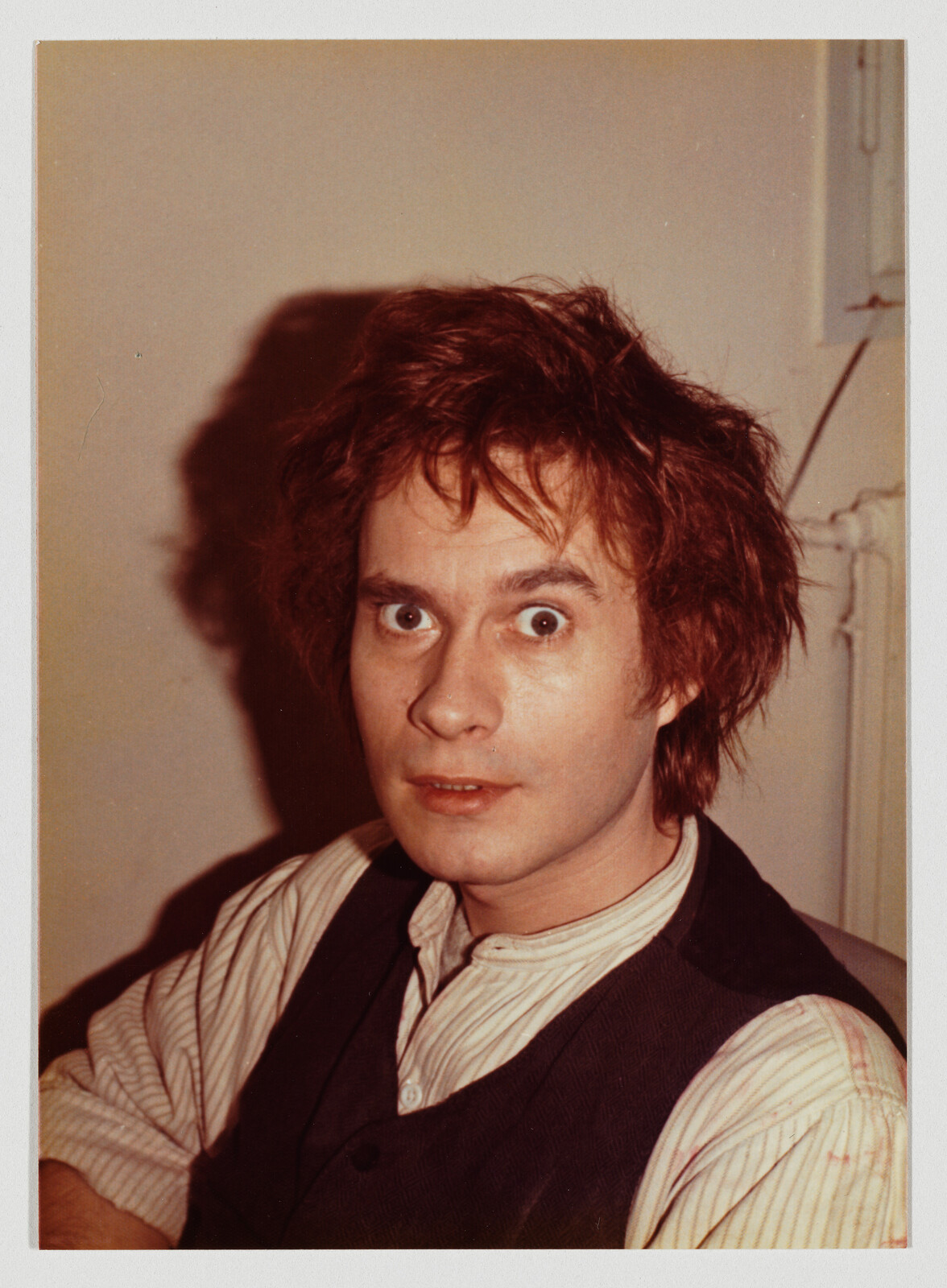 A young man with messy hair and wide eyes looks surprised while wearing a striped shirt and vest.