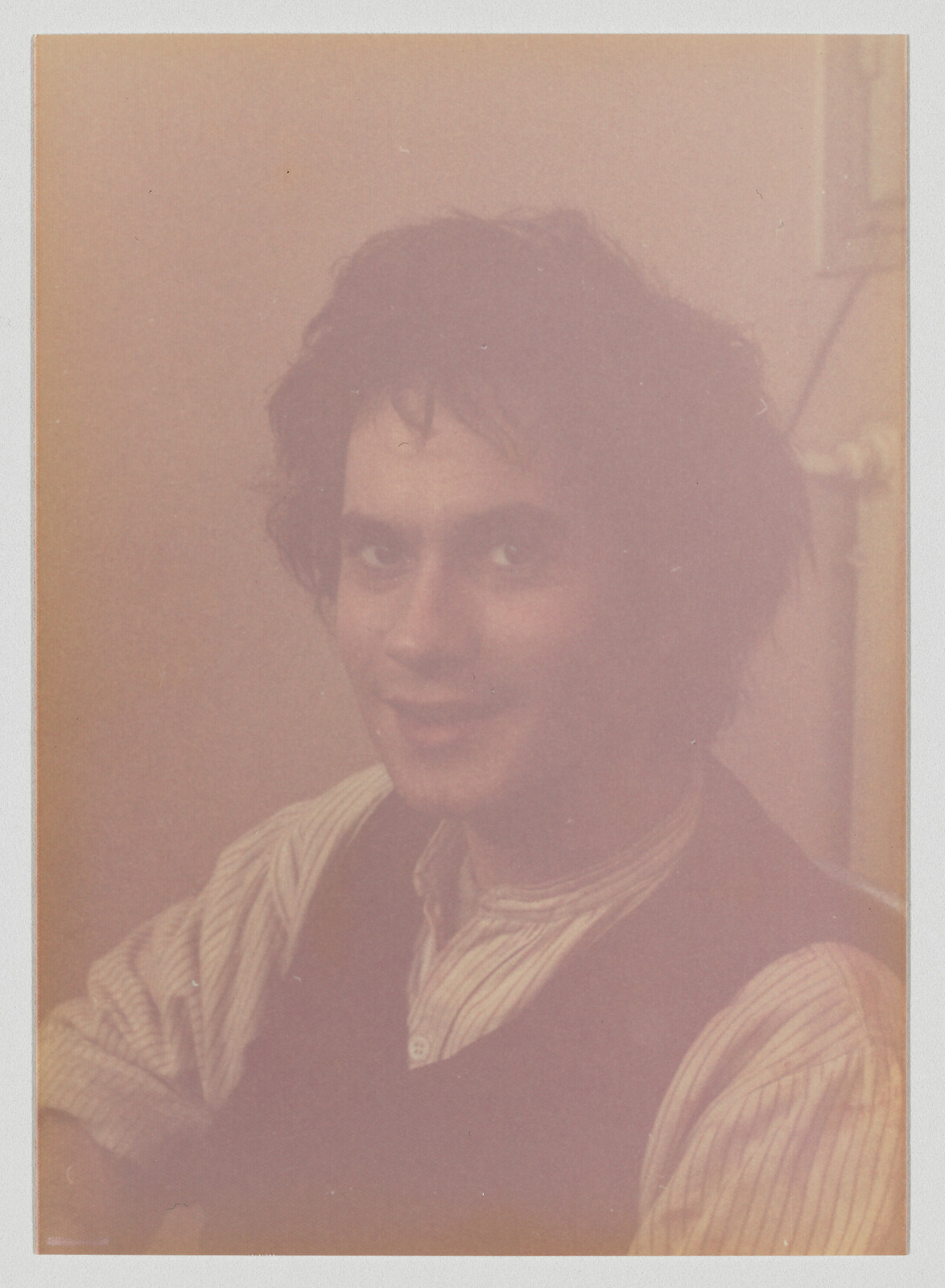 A young man with messy hair smiles softly while wearing a striped shirt and dark vest.